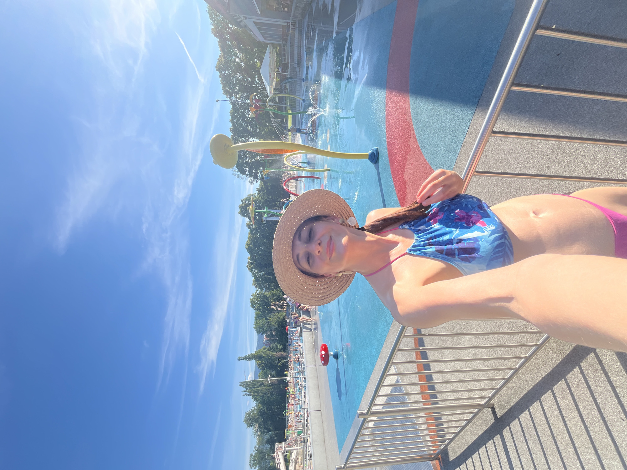 A woman wearing a straw hat and a blue and pink patterned bikini is taking a selfie in a public splash pad on a sunny day. She is standing behind a silver metal railing, with a bright blue splash pad area in front of her. Several water fountains and structures are visible in the background, along with trees and buildings. The sky is a clear blue with some wispy clouds.