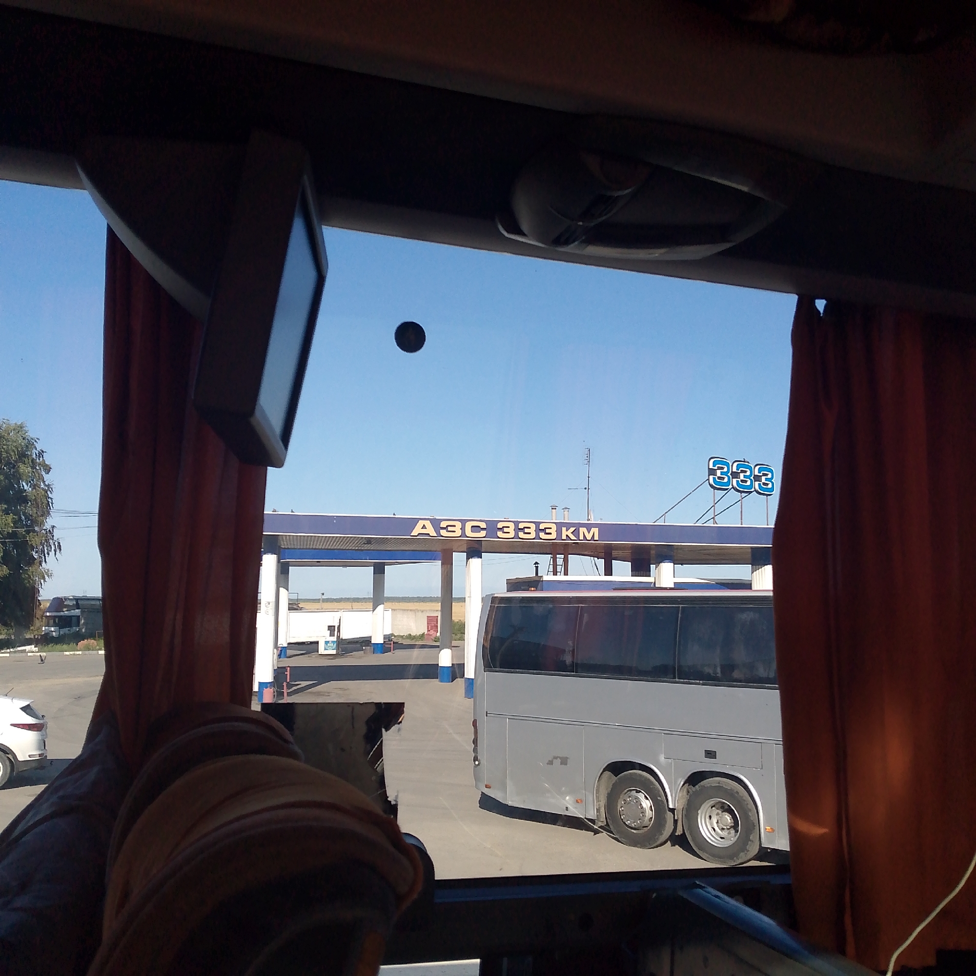 The image captures a view from inside a vehicle, looking out through a window at a gas station. The gas station has a blue canopy with the yellow text "АЗС 333KM" prominently displayed, and a smaller blue sign with "333" in white text. A large, silver bus is parked at the gas station, with its rear visible. In the foreground, part of the interior of the vehicle, including orange curtains and a dark dashboard, is visible. To the left, a white car is parked, and a tree stands behind it. The sky is clear and blue. The scene appears to be a sunny day. While the image itself doesn't explicitly state the location, the gas station signage "АЗС 333KM" suggests it could be along a highway route marked by kilometer markers, and the presence of a bus indicates a transportation hub or route. Without further information, pinpointing the exact location in Domodedovo, Russia, is not possible from the image alone.