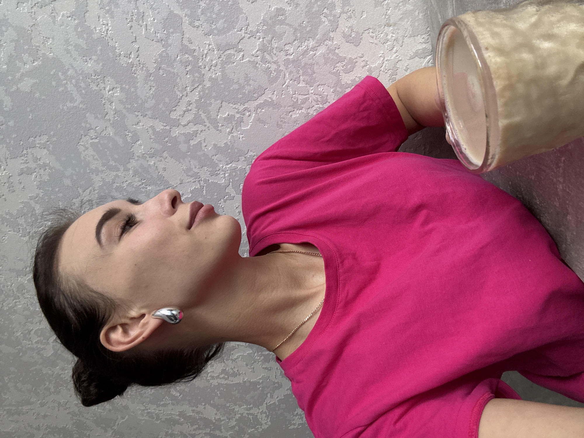 The image features a young woman in a relaxed, reclined position, looking upwards and to her left. She has dark hair pulled back, well-defined eyebrows, and her eyes appear closed or heavily lidded, suggesting a tranquil or restful state. Her lips are slightly parted. She is wearing a bright fuchsia pink short-sleeved t-shirt and a delicate gold-toned chain necklace. A notable accessory is the large, sculptural, silver-toned earring with a smooth, organic shape on her right earlobe.
To her right, a clear glass jar or large mug, holding a light brown, foamy beverage (possibly a smoothie or milkshake), is visible. The woman appears to be holding it or resting it nearby. The background is a light grey wall with a textured, subtly patterned wallpaper. The lighting is soft and consistent, indicating an indoor setting, likely domestic. No text is visible in the image. The scene conveys a sense of calm and quiet.