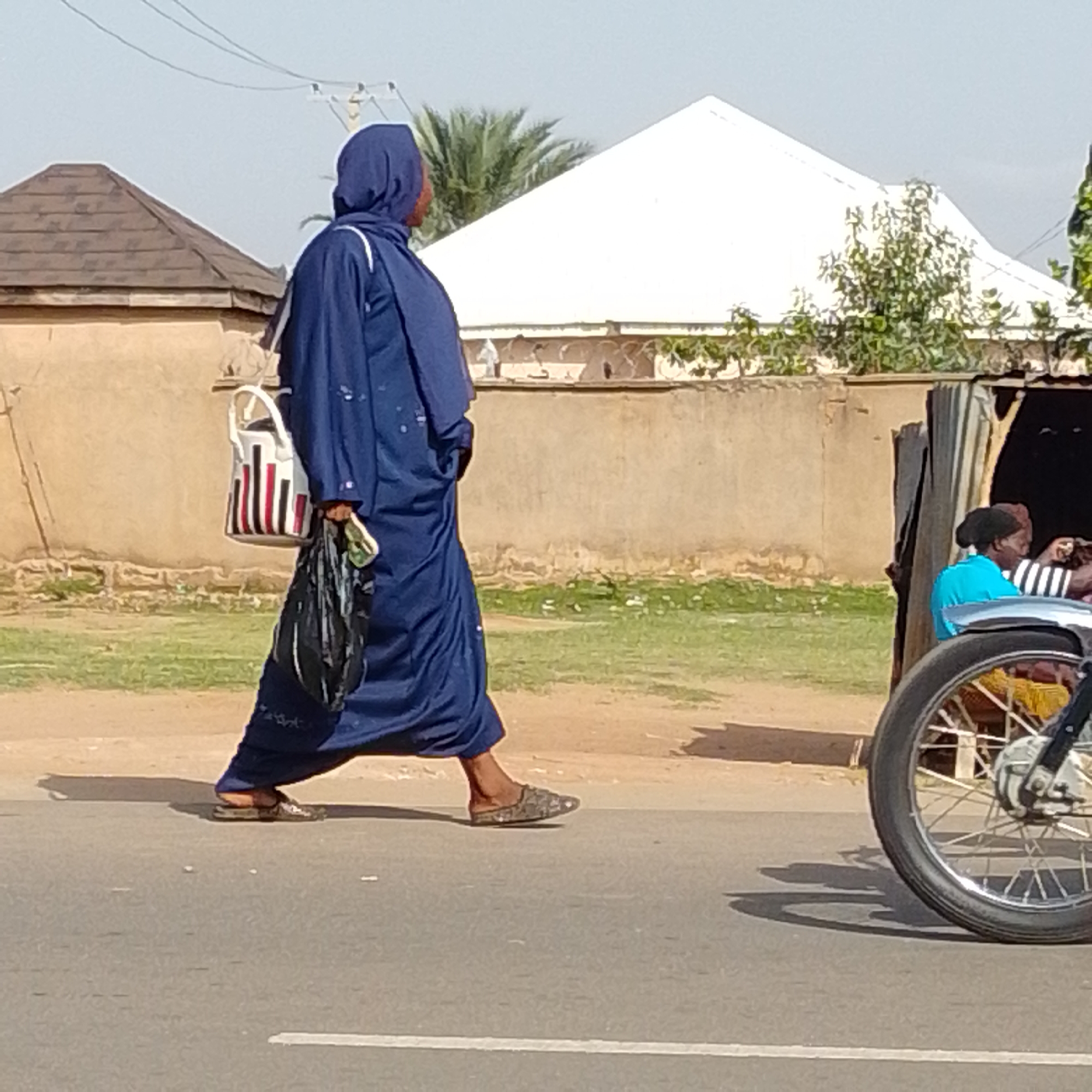 In the city of Gombe, Nigeria, a woman in a blue abaya and hijab walks along the side of a paved road. She carries a black plastic bag in her right hand and a white and red striped bag slung over her left shoulder. In the background, a light brown wall separates the road from a grassy area. Beyond the wall, a white, peaked roof structure and a brown, gabled roof structure are visible. To the right of the frame, a motorcycle wheel is prominent, and a small stall or shelter with people inside is partially visible. The sky is clear and bright.