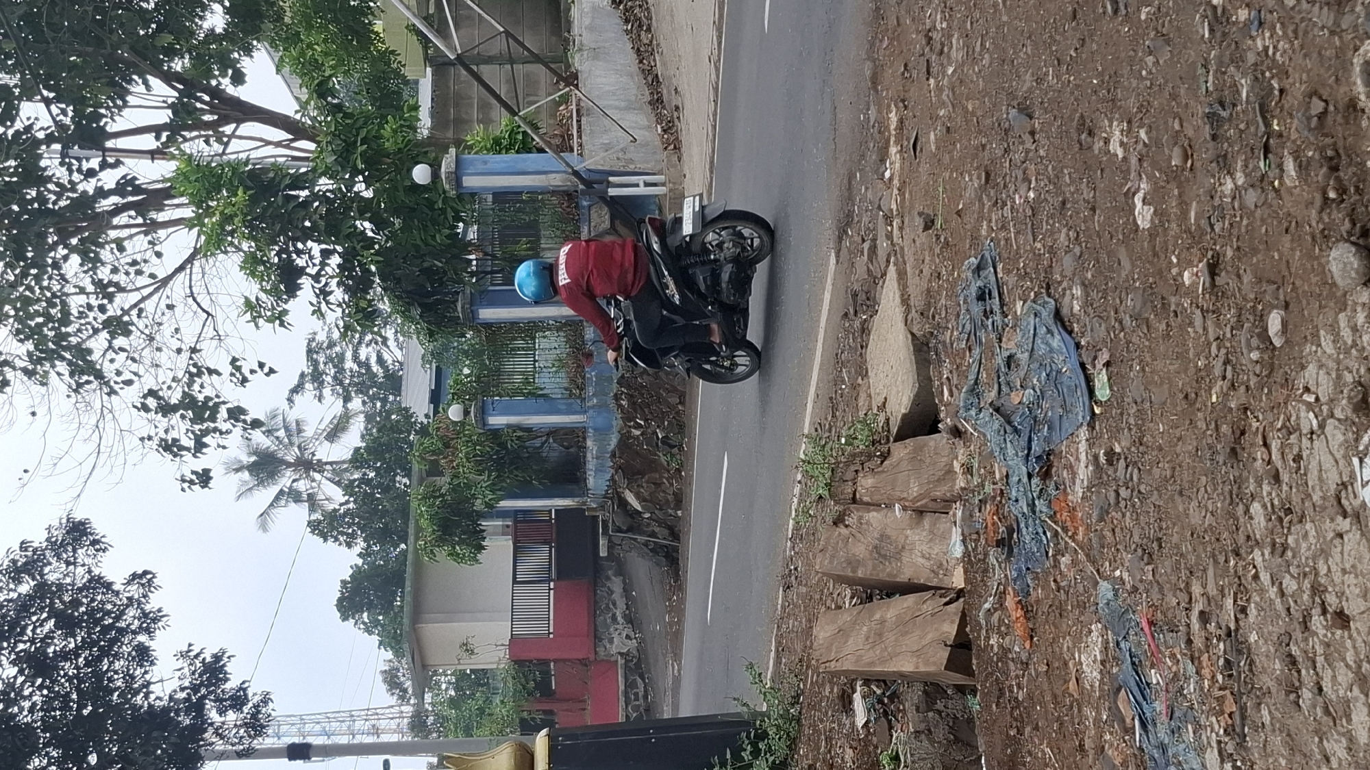 A person wearing a red shirt and a blue helmet rides a black motorcycle on a paved road in Cilaku, Indonesia. The person is viewed from behind and to the right, with the motorcycle moving from left to right. The road has a white line marking its center. To the left of the road, there is a dirt area with some debris, including blue plastic sheeting and pieces of wood. In the background, there is a building with red and white walls, a blue fence with white pillars, and trees. A tall tower is visible in the distance. The sky is overcast.
