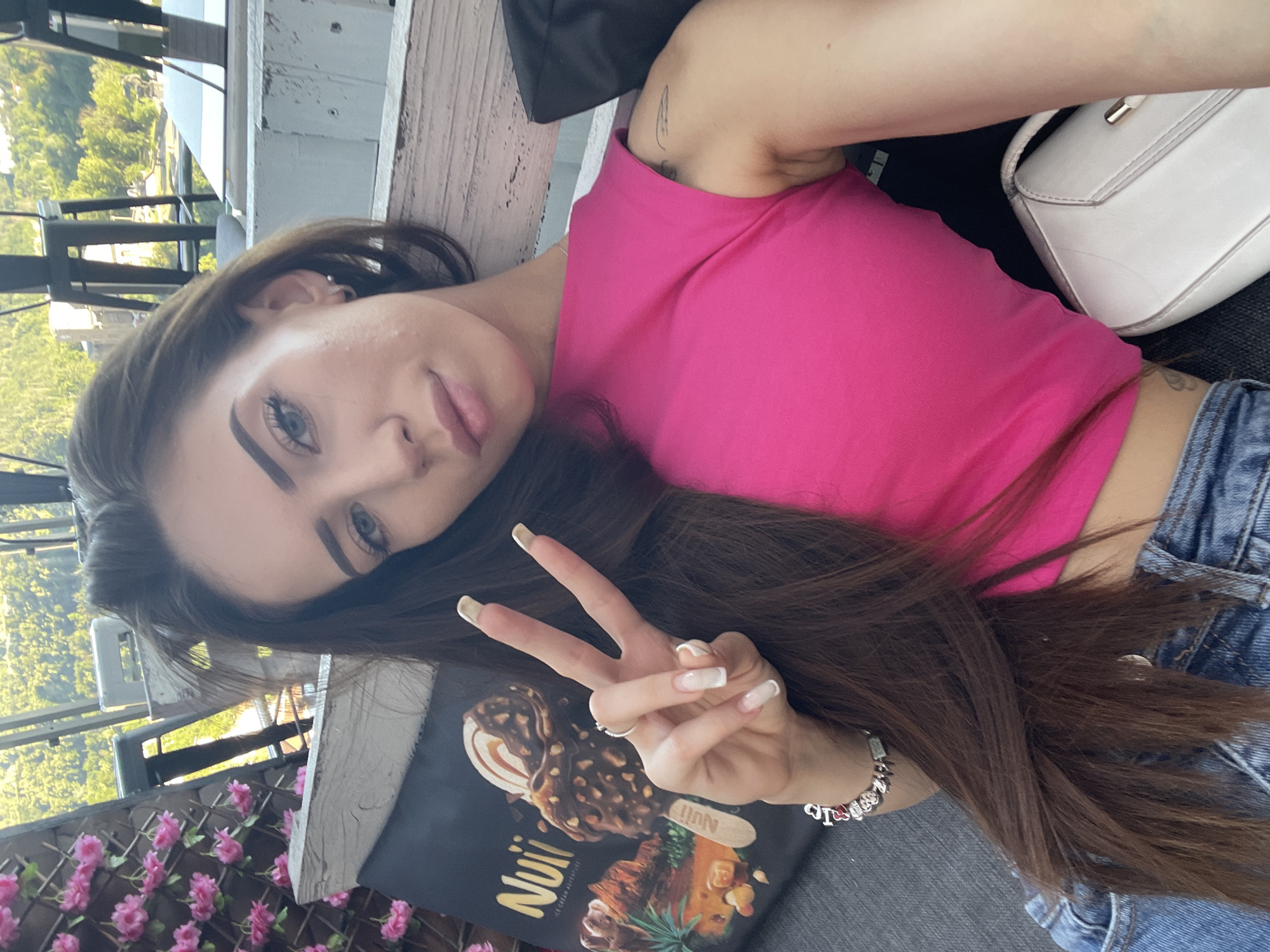 A young woman with long, dark brown hair is seated outdoors, making a peace sign with her right hand. She is wearing a bright pink crop top and blue jeans. In the background, a large black sign with an image of an ice cream bar and the word "Nuii" is visible. She is also wearing a silver bracelet with charms and a ring on her left hand. A white purse rests beside her. The setting appears to be a restaurant or bar with a view of trees and buildings in the distance.