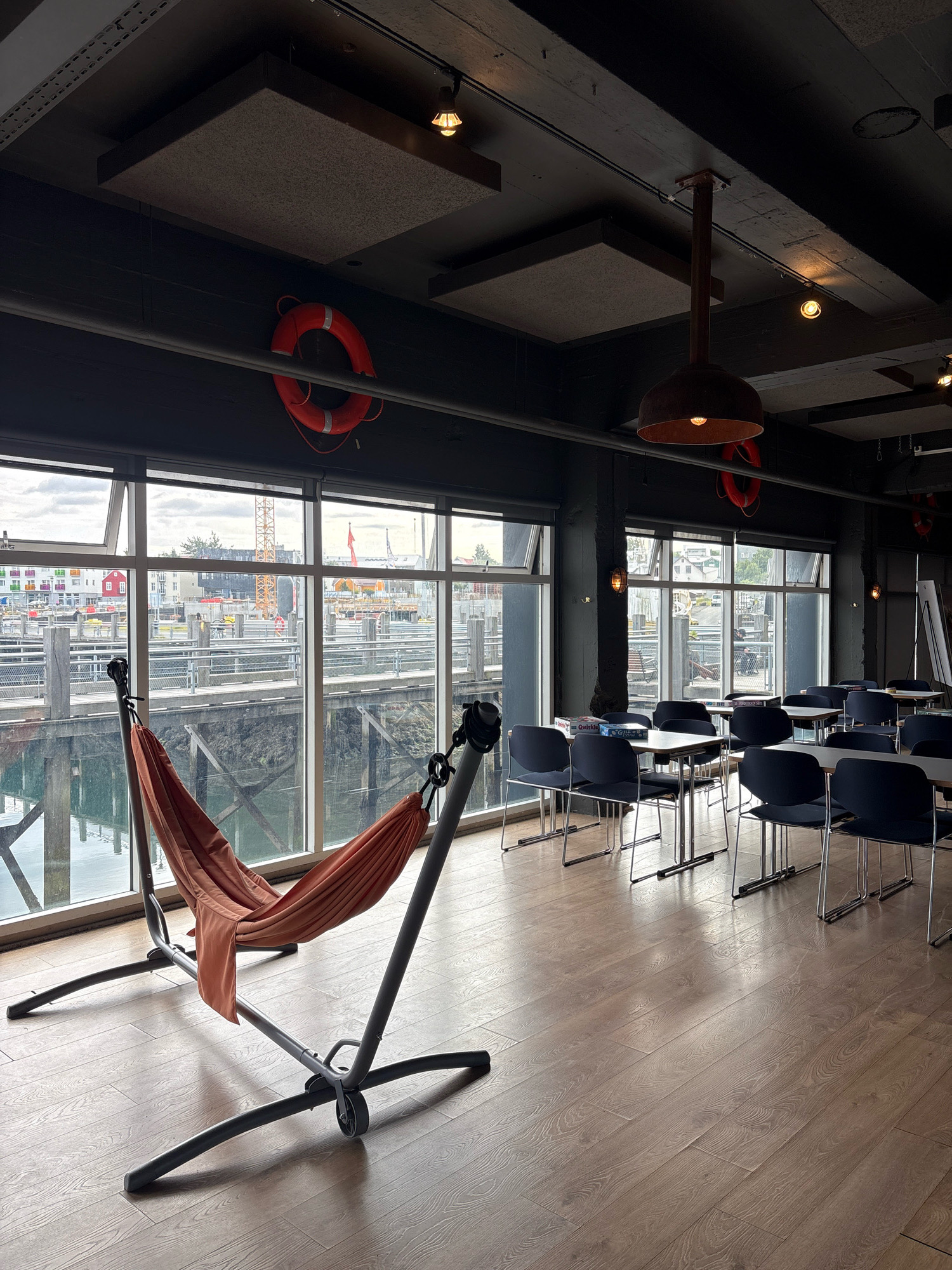 This image depicts the interior of a cafe or common area with large windows overlooking a harbor in Reykjavik, Iceland. In the foreground, a coral-colored hammock is suspended from a dark gray metal stand. The hammock is empty and draped loosely. Behind the hammock, a row of dark blue chairs surrounds several light-colored rectangular tables. More tables and chairs are visible further back in the room.
The large windows offer a view of the harbor, with piers, railings, and boats. In the distance, buildings and a construction crane are visible, characteristic of a port city. The ceiling is dark and features exposed beams, acoustic panels, and industrial-style lighting fixtures. Two orange life preservers are mounted on the wall above the windows. The floor is made of light-colored wooden planks. The overall atmosphere suggests a relaxed, indoor space with an industrial or nautical aesthetic.