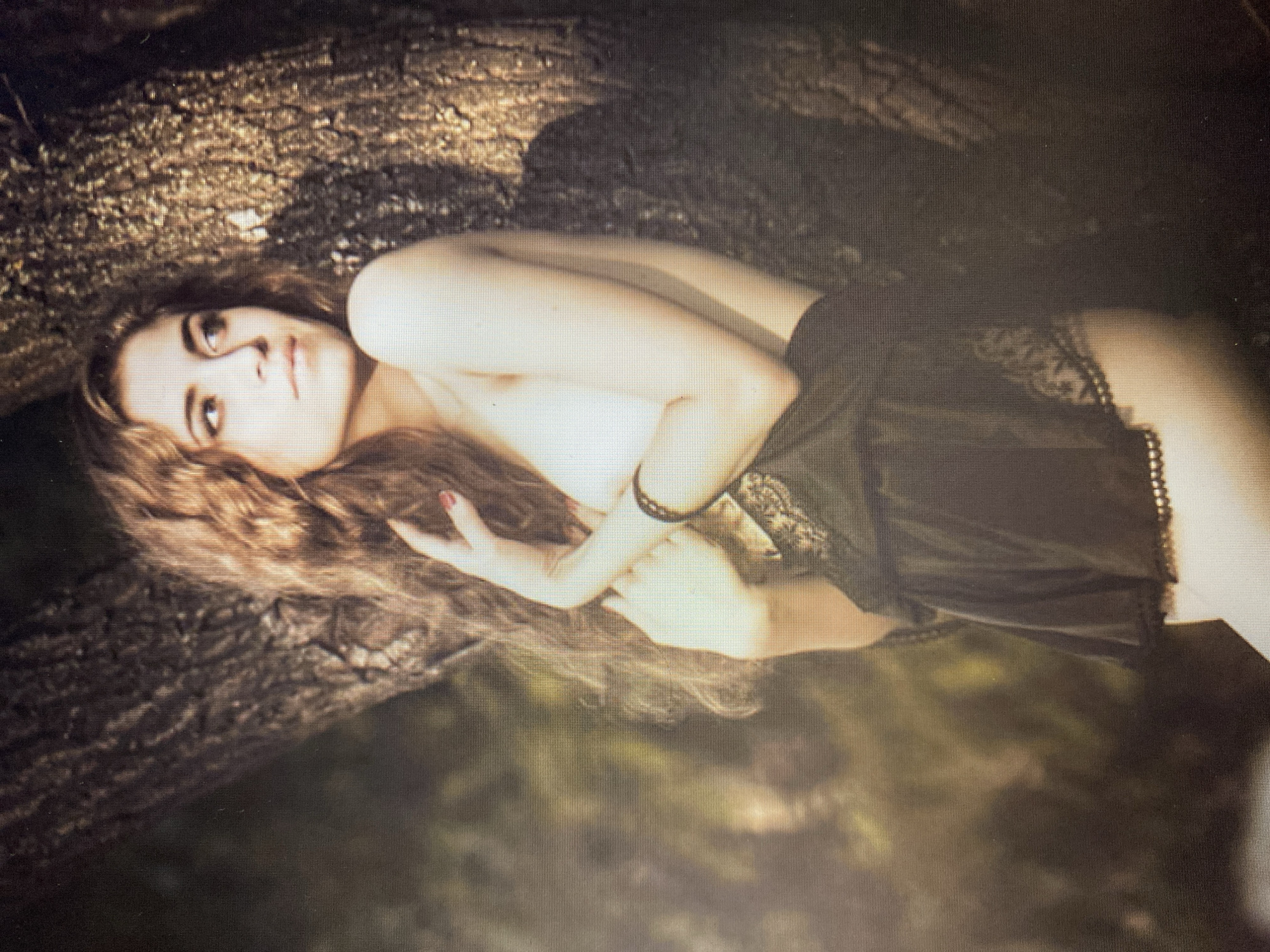 A young woman with long, wavy brown hair is leaning against a tree. She is wearing a black lace top and skirt. Her left arm is bent, and her hand is resting on her chest. Her right arm is behind her head. The background is a blur of green and brown, suggesting an outdoor setting. The lighting is soft and natural, casting a warm glow on the woman's skin.