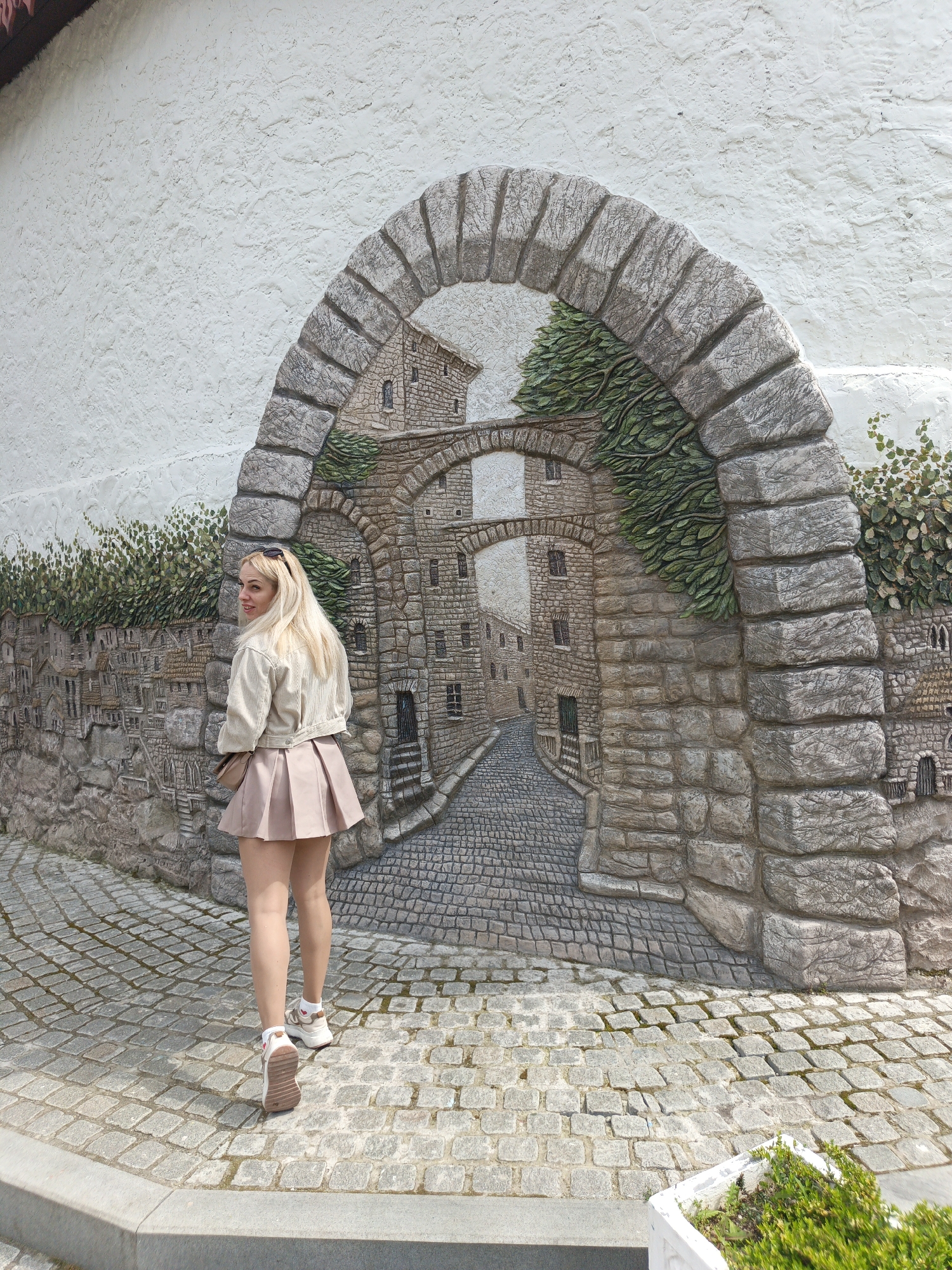 A full outdoor shot depicts a blonde woman in a short pink skirt and light jacket, walking away from the viewer and slightly turned to her left, in front of a stone archway mural. The mural, painted on a textured white wall, portrays a receding cobblestone street and building facades. The setting appears to be on a cobblestone paved surface, possibly in a city within the Алупкинский городской совет region of Ukraine, although no specific landmarks identify the exact location.