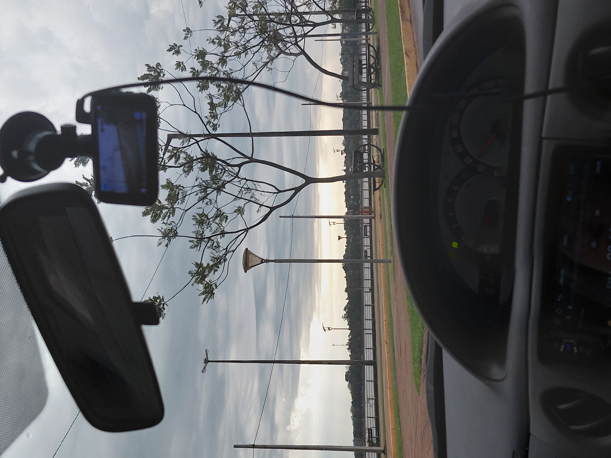 The image is a point-of-view shot from inside a car, looking forward through the windshield. A rearview mirror is visible at the top of the frame, and a dashcam is mounted to the windshield below it. The car is parked on a street adjacent to a waterfront area with benches and trees. In the background, a body of water, possibly a river, is visible, with a tree-lined shore on the opposite side. The sky is overcast with a hint of sunlight breaking through. The dashboard of the car is visible in the foreground, with a speedometer and other gauges. The scene appears to be a public park or promenade, likely in the city of Encarnación, Paraguay, given the context. There are no people visible in the image.