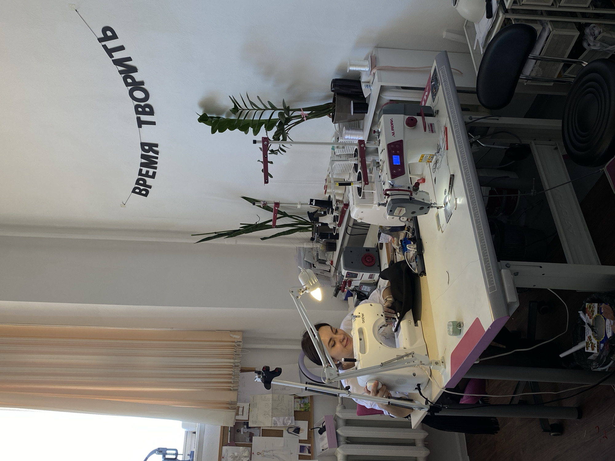 A woman is seated at a sewing machine in a bright room, likely a tailor's workshop. She is focused on her sewing task, using a white Brother sewing machine. The room contains multiple sewing machines on a long table. The workshop has plants in pots placed amongst the machines. There is decorative text reading "Время Творить" (Time to Create) hanging on the wall. A window with blinds lets in natural light. The location is possibly Izhevsk, Russia.