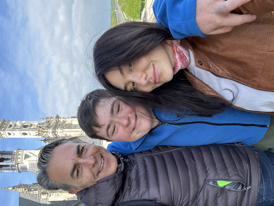 Three people are standing close together outdoors, taking a selfie. The man on the left, wearing a black puffy vest over a blue shirt and jeans, has his arm around the middle person, a young man wearing a blue zip-up jacket. The woman on the right is wearing a tan leather jacket over a white t-shirt, and a pink patterned scarf around her neck. She is looking at the camera. The background is a white stone building with a tall tower and a blue sky with white clouds. This location is the Château de Chambord, located at coordinates (latitude: 47.616, longitude: 1.518) in Chambord, France.