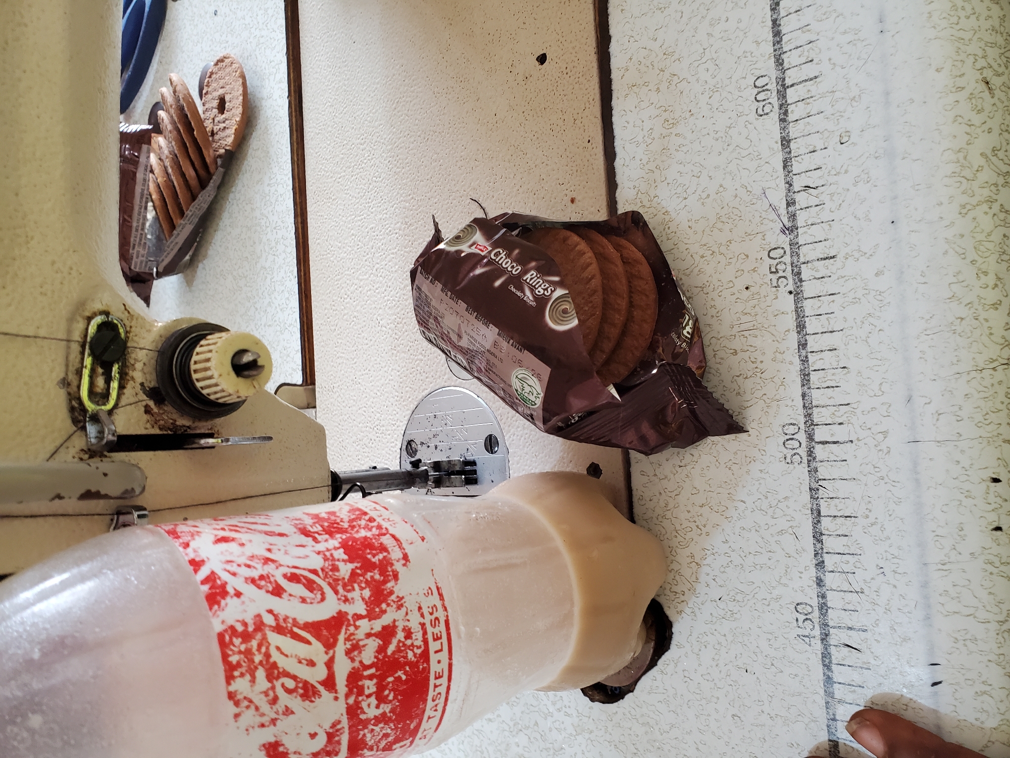 The image presents a close-up, high-angle view of a work surface, likely a sewing table, featuring various objects and a human hand. In the foreground, a light-colored plastic bottle with a distinct red and white patterned label, reminiscent of Coca-Cola branding, rests on the table, its contents appearing milky white. Part of the text "TASTE LESS" is visible on the bottle. To the left, a portion of a cream-colored sewing machine is prominent, with its mechanical components and a storage compartment where several "Choco Rings Chocolaty Biscuits" are stacked. Another open packet of these same biscuits is positioned centrally on the table, revealing more of the brown, ring-shaped cookies.
The table surface itself is a textured, light beige or off-white material, imprinted with a ruler marked with numbers "450", "500", "550", and "600". A human thumb and part of a finger are visible in the bottom right corner, resting near the "450" mark on the ruler. The scene suggests a brief break from work at a sewing station, with snacks and a drink readily available. The biscuit packet text includes "Choco Rings Chocolaty Biscuits", "BATCH NO: MFG DATE: BEST BEFORE: MIEUX AVANT", "P20707254", "BB105/26", and "PARDEE FOODS NIGERIA LTD", indicating the origin of the product. No specific time of day or weather is discernible, but the setting is clearly indoors.
