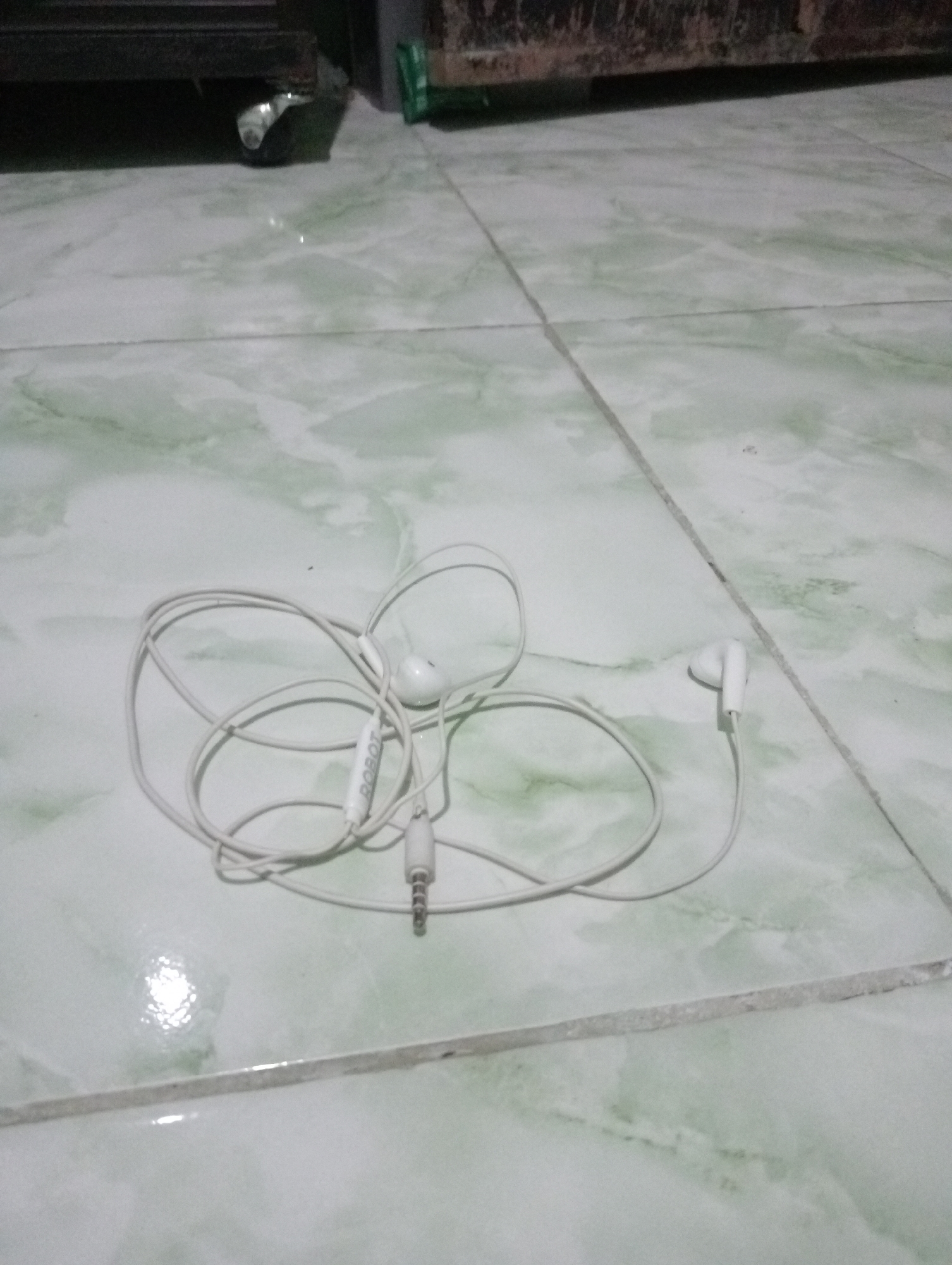A tangled pair of white wired earbuds rests on a pale green and white marbled tile floor. The earbuds have white in-ear buds and a white cord with a visible "ROBOT" logo on the Y-split. The 3.5mm audio jack is visible on the right side of the image. The background shows the bottom of a dark piece of furniture with a visible wheel. The overall lighting is dim.