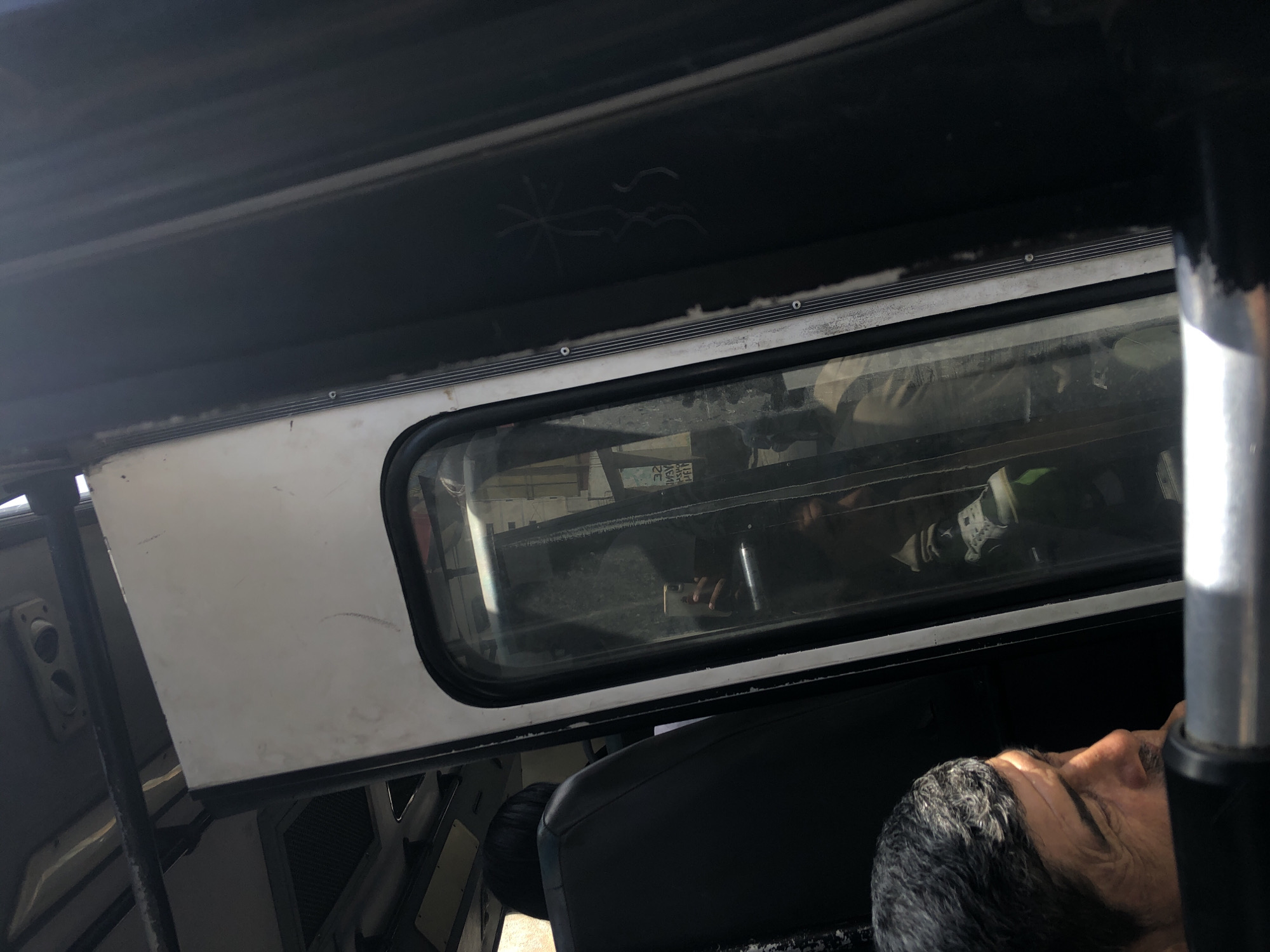 The scene appears to be the interior of a vehicle, possibly a bus or truck, observed from the perspective of someone inside. There's a window showing a reflection of the interior space, including a person holding a phone. A man's head and part of his shoulder are visible in the foreground, suggesting he is seated in the vehicle. The vehicle's interior is utilitarian and somewhat worn, featuring metal framing and a black roof liner. The environment reflected in the window is blurry but suggests an urban setting. There is no definitive information about Porlamar, Venezuela.