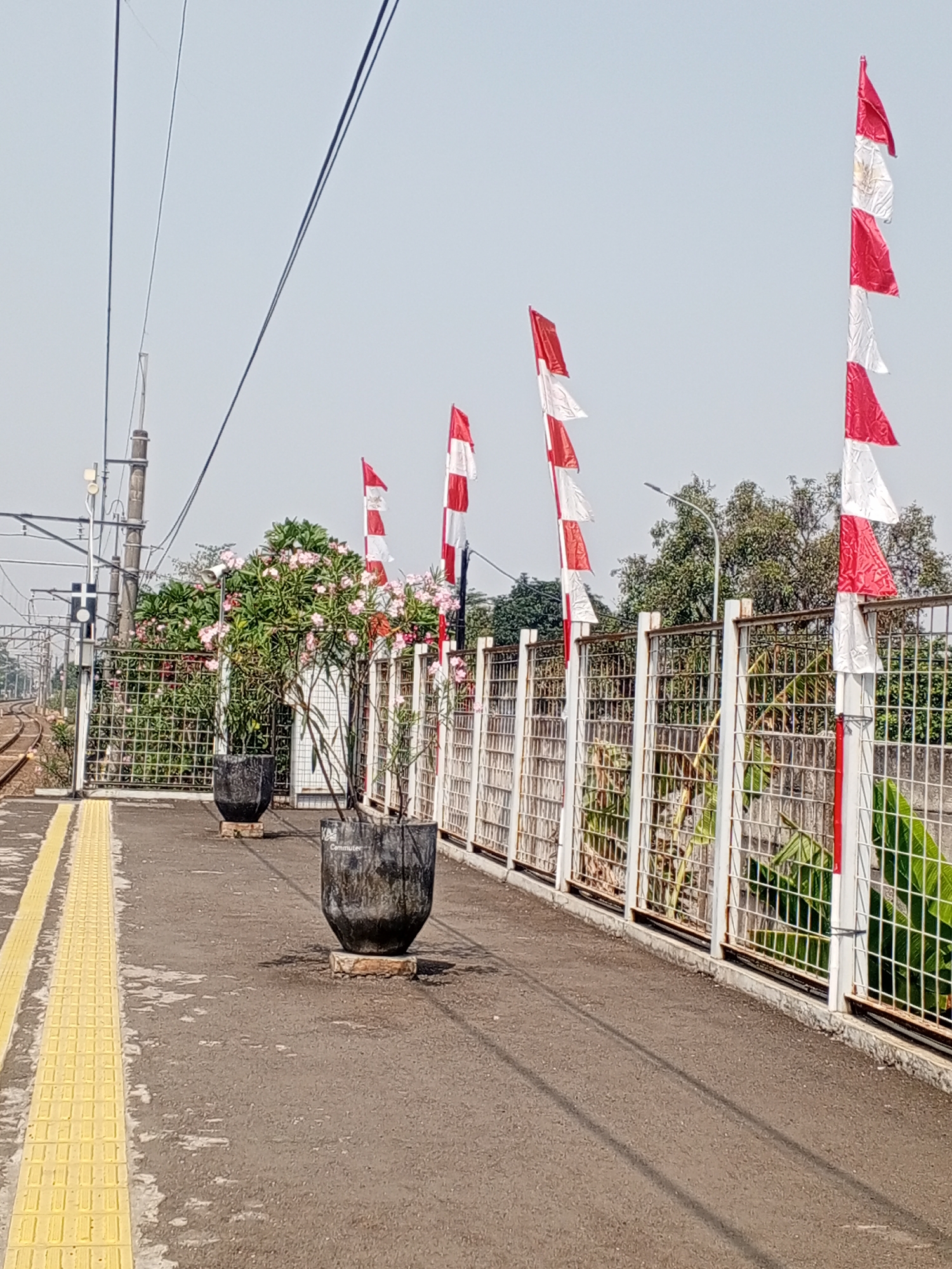 The image shows a view of a train station platform in Depok, Indonesia. The platform is paved with asphalt, with a yellow tactile paving strip along the edge. A white metal fence runs along the side of the platform, separating it from vegetation and trees beyond. Several Indonesian flags, red and white, are mounted on poles along the fence, fluttering in the breeze. Large black planters containing pink flowering plants are placed on the platform. Overhead, power lines crisscross the sky, supported by utility poles. In the background, train tracks curve away from the station. The sky is clear and bright.