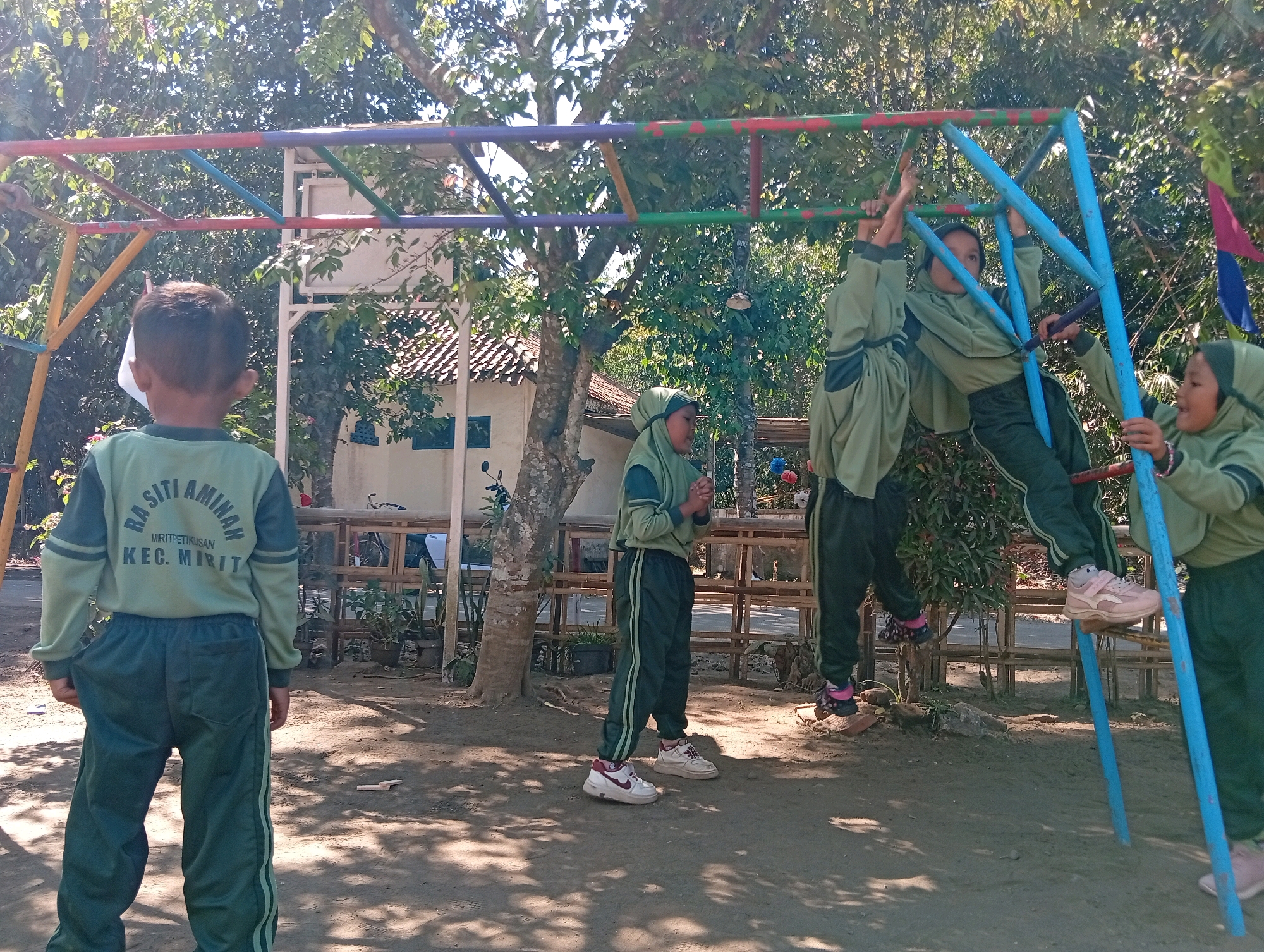 In Miritpetikusan, Indonesia, on a sunny day, a group of children are playing on a colorful metal jungle gym. The jungle gym is blue, red, yellow, and purple. Four children are visible, all wearing matching green school uniforms with darker green accents and white trim. One child, seen from the back, stands on the sandy ground, facing the jungle gym. Another child, wearing a hijab, is on the jungle gym, holding onto a blue pole. A third child, also in a hijab, is climbing on the jungle gym, holding onto a horizontal bar. The fourth child, in a hijab, is further to the right, also interacting with the jungle gym. The background features lush green trees and a white building with a tiled roof, typical of a school or residential area. The ground is covered in sand and dappled with sunlight and shadows from the trees. The children's focused expressions and active poses indicate engagement in playful activity.