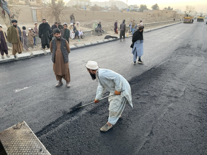 This image depicts an active road paving operation in Kandahar, Afghanistan, under an overcast or late afternoon sky. In the foreground, a bearded man wearing a white turban, a light-colored shalwar kameez, and orange gloves is bent over, using a long-handled rake to spread dark, freshly laid asphalt. A portion of a diamond-plate metal surface, possibly from an asphalt paver, is visible at the bottom left.
To the left of the active worker, a man with a short beard, dressed in a brown shalwar kameez and dark vest, stands looking directly forward. To his right, another man wearing a black shawl over a light blue shalwar kameez stands with his back partially to the viewer, observing the ongoing roadwork.
Along a raised concrete barrier or curb to the left side of the road, a crowd of approximately 10-15 men and boys, mostly in traditional Afghan attire, are gathered as observers; some are standing, while others are seated.
The newly paved road extends into the mid-ground and background, where a yellow road roller is visible compacting the asphalt. Another yellow piece of heavy machinery is also present further down the road. The environment includes low-rise buildings along the roadside and distant mountains under a hazy sky.