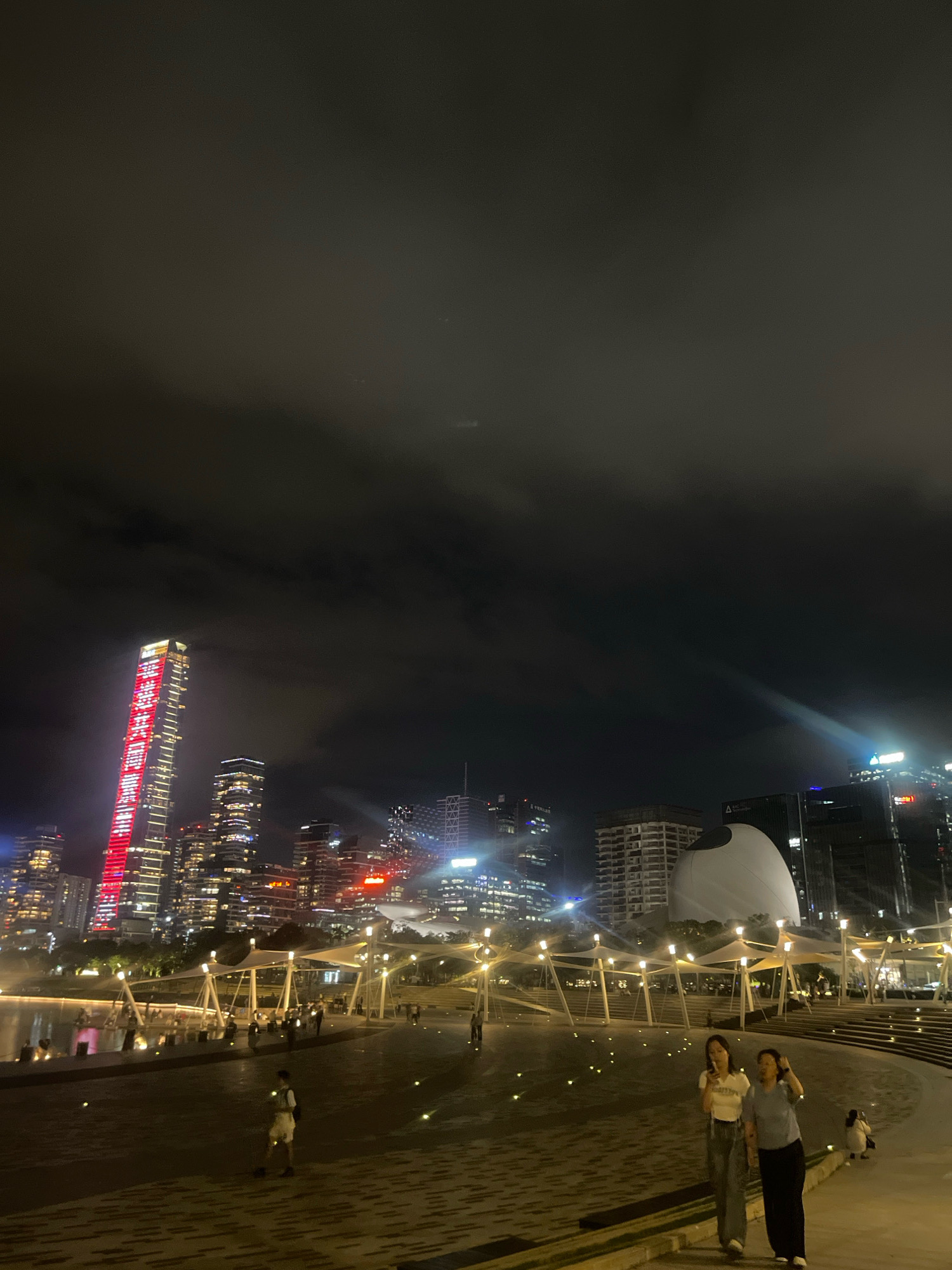 This is a nighttime photograph of a cityscape in Nanshan District, China. In the background, a cluster of illuminated skyscrapers dominates the skyline. The tallest building on the left is adorned with red lights forming characters. In the foreground, a paved area with scattered lights leads to a body of water. Several people are visible, with two women walking towards the viewer in the center of the frame. The environment appears to be a public space or park, possibly a waterfront promenade, with architectural features like a large, white, dome-shaped structure and a canopy-covered walkway. The sky is dark and partially obscured by clouds.