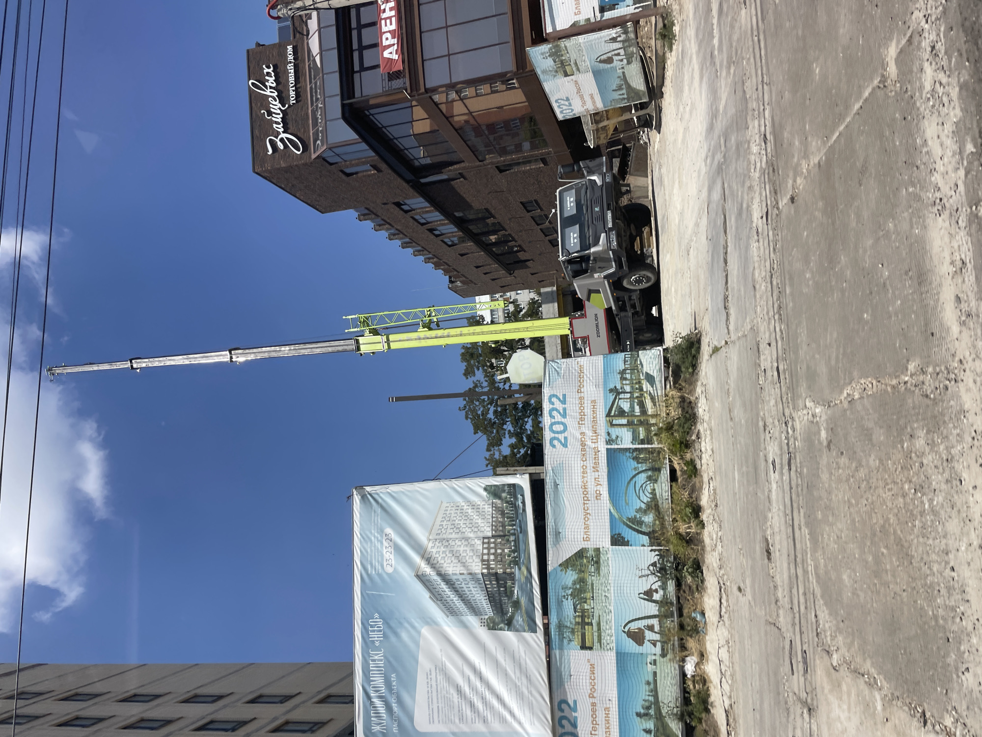 The image captures a street view in Stavropol, Russia, likely near a construction site. A crane, identified by the "ZOOMLLION" logo on the truck it's mounted on, is visible, its boom extending upwards. In the background, a modern brick building with large windows displays a sign with stylized Cyrillic script that reads "Зайцевых" and "ТОРГОВЫЙ ДОМ," indicating a department store. Another sign on the building reads "АРЕН" suggesting commercial space is available for rent.
In the foreground, banners are displayed. One large banner advertises a residential complex named "НЕБО" (Sky), featuring an architectural rendering of a multi-story building and project details in Cyrillic. Below this, smaller banners showcase renderings of a park and public spaces, with the year "2022" and text "Благоустройство сквера 'Героев России' пр. ул. Ивана Цапакина" (Beautification of the square 'Heroes of Russia' on Ivan Tsapakin street). This confirms the location as Stavropol, specifically near Ivan Tsapakin street.
A truck, likely a mobile crane, is parked on a damaged, uneven paved surface. The area appears to be under development or has recently undergone some work, with debris and cracks visible on the road. The sky is blue with some clouds. No people are clearly visible in the image.