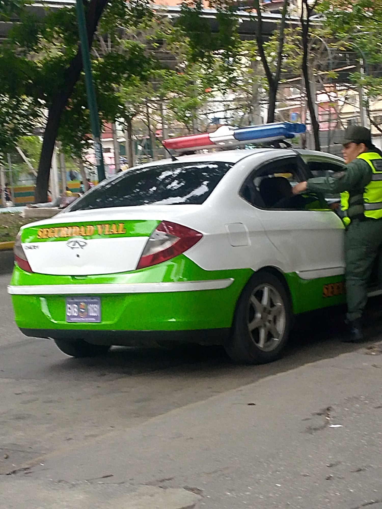 The image shows a white and green Chery Arauca Seguridad Vial vehicle with a red and blue light bar on top, parked on a city street. The vehicle has the words "SEGURIDAD VIAL" written on the back in yellow letters. A person wearing a green uniform and a yellow vest is opening the driver-side door. The license plate is visible, reading GNB 1061. Trees and buildings are visible in the background, suggesting an urban environment in Caracas, Venezuela.