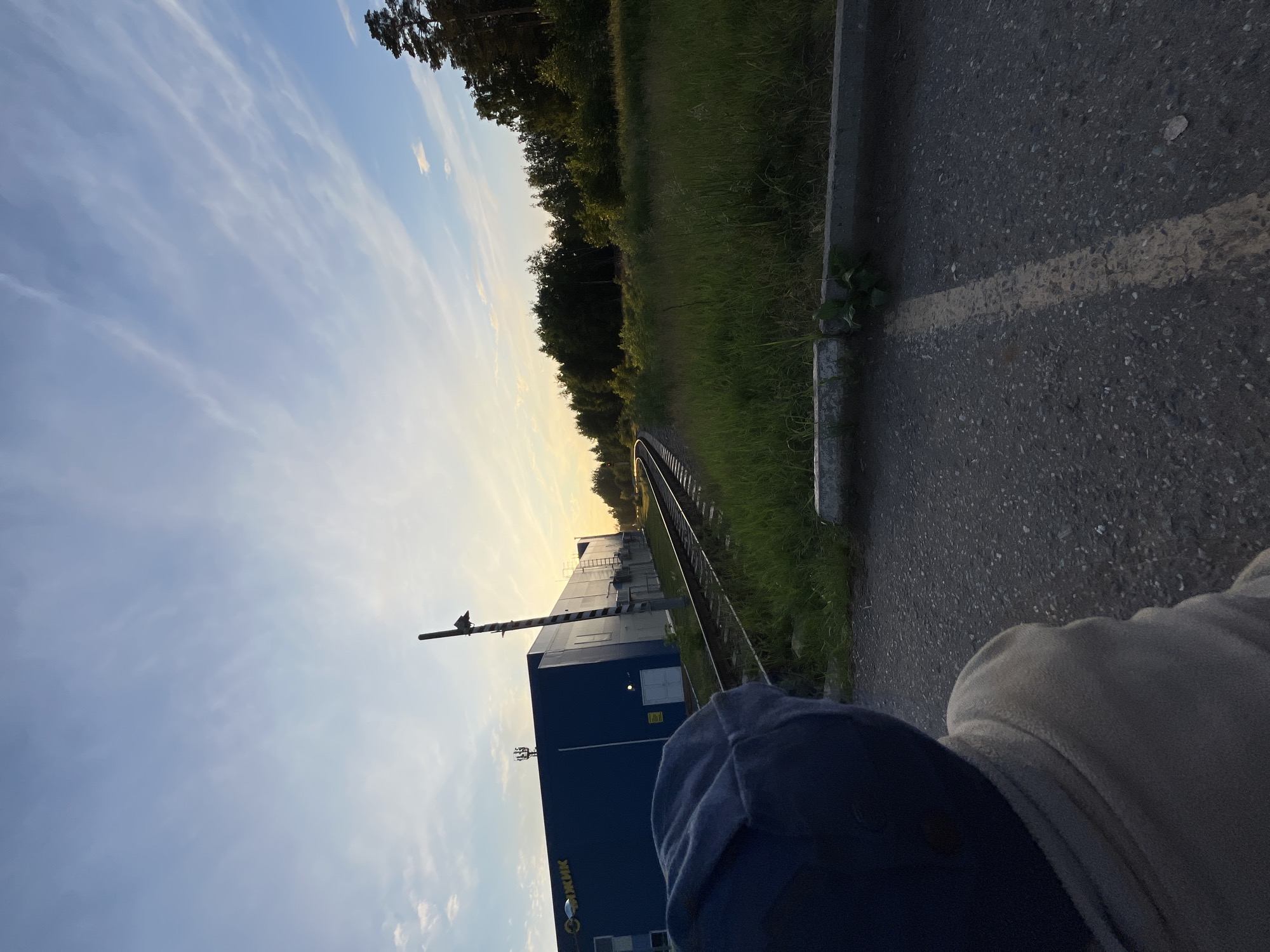 A rear view of a person wearing a blue hooded jacket and a blue cap is facing away from the camera, looking towards a railway track. To the left, a long, blue industrial building with the word "Чайковский" in white text is visible. The building has several windows, doors, and antennae on its roof. A tall pole with a light and striped pattern stands beside the railway tracks. The tracks curve to the left and disappear into a grassy field bordered by trees. The sky above is a pale blue with wispy white clouds, illuminated by the warm glow of the setting sun. The ground in the foreground is asphalt with a faded yellow line.