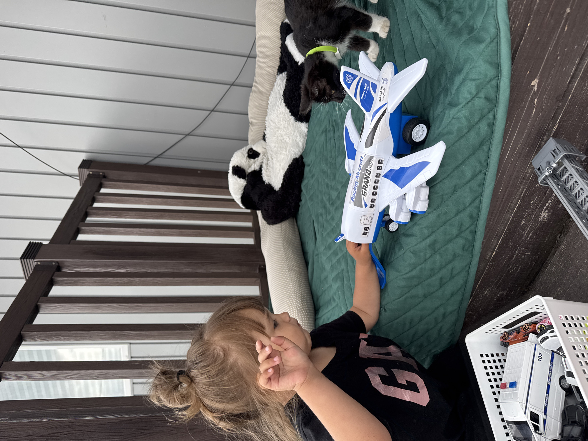 A young Caucasian girl with blonde hair in a messy bun is sitting on a green mat outdoors, playing with a white and blue toy airplane. She is wearing a black t-shirt with pink lettering. To her right, a black and white kitten with a green collar is sniffing the airplane. In the background, a large stuffed panda bear is lying on a beige pillow. The scene appears to be on a wooden deck or balcony, with a wooden railing and a white-paneled wall behind. To the left of the girl is a white plastic basket filled with toy cars, including a police van. A metal toy construction vehicle is also visible on the wooden surface in the foreground. The general impression is that this is a residential area in Kempelevo, Russia, likely a backyard or patio, where a child is engaged in playtime with pets and toys.