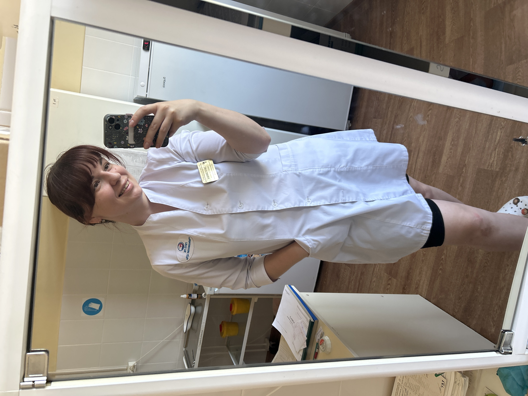 A woman stands in front of a mirror in a medical office in Фоки, Russia, holding a phone and taking a selfie. She is wearing a white medical coat with a name tag on the left side. The office environment is visible in the mirror's reflection, showing cabinets, medical supplies, and tiled walls.