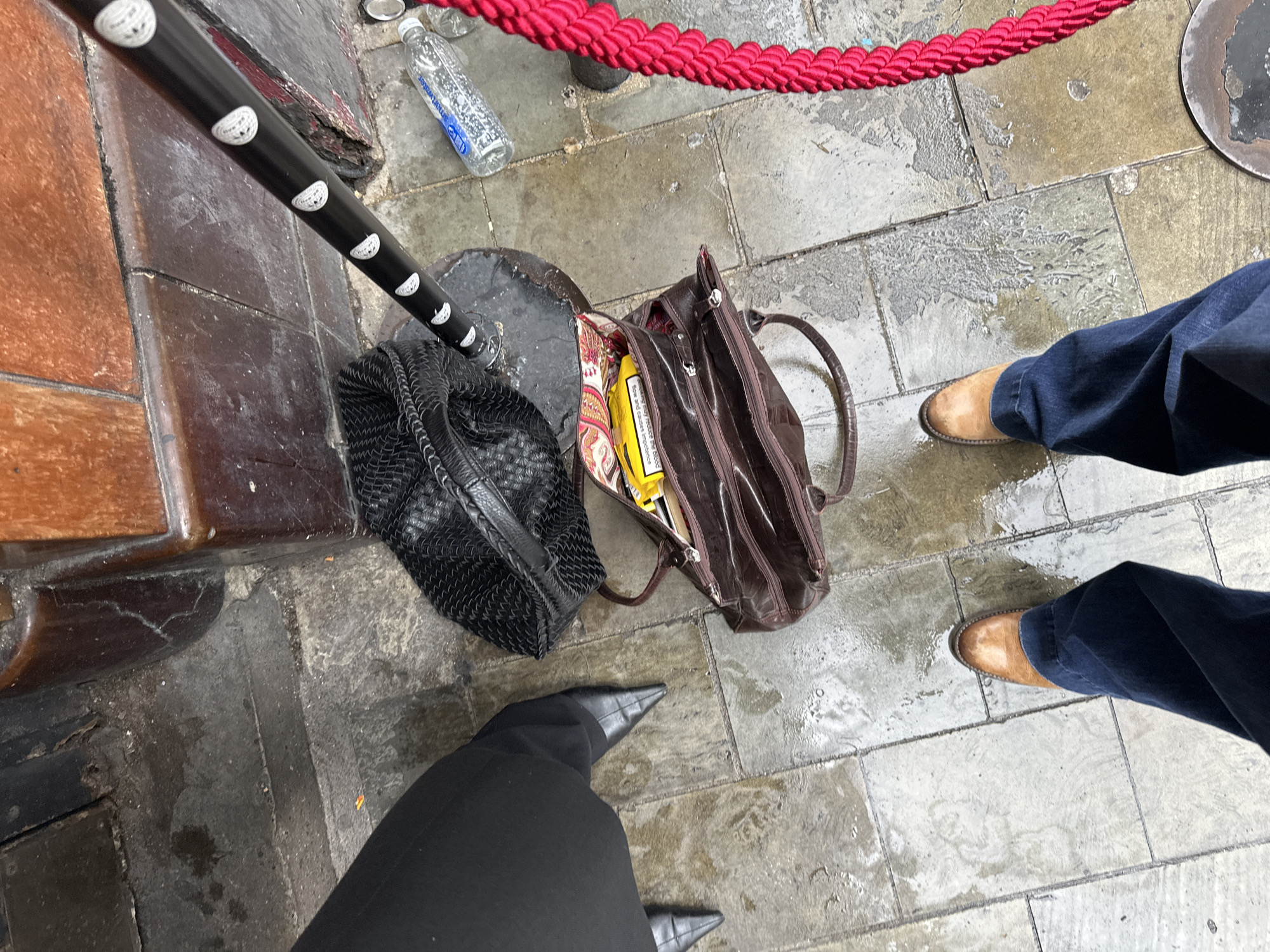 This overhead image depicts a wet, grey and brown tiled pavement scene in London, United Kingdom, showing the ground level adjacent to a building facade. Two individuals' lower legs and feet are visible. On the left, a person wears black trousers and black, pointed-toe, high-heeled shoes. In the foreground, another person wears dark blue jeans and tan, pointed-toe boots.
Multiple objects are present on the damp pavement. A black woven-textured handbag is partially visible on the left, positioned beneath a black stanchion-like pole. This pole runs diagonally across the frame from top-right to mid-left and features several white circular emblems with crescent moon designs. To the right of the pole, a transparent "smartwater" plastic bottle, partially filled with liquid, lies on its side. Centrally located, an open brown leather handbag contains visible items, including a yellow package with partially legible text, "WARNING: Smoking seriously harms you and others around you. Smoking causes...", identifying it as a cigarette pack bearing a UK/EU health warning. A patterned fabric, possibly a scarf, is also visible inside the open bag. On the far right, a thick, bright red braided rope, likely part of a crowd control barrier, extends from a metal base on the ground. The background features a dark brown/orange tiled or paneled wall, possibly the lower section of a building facade, exhibiting several cracks. The pavement shows consistent wetness across its surface.