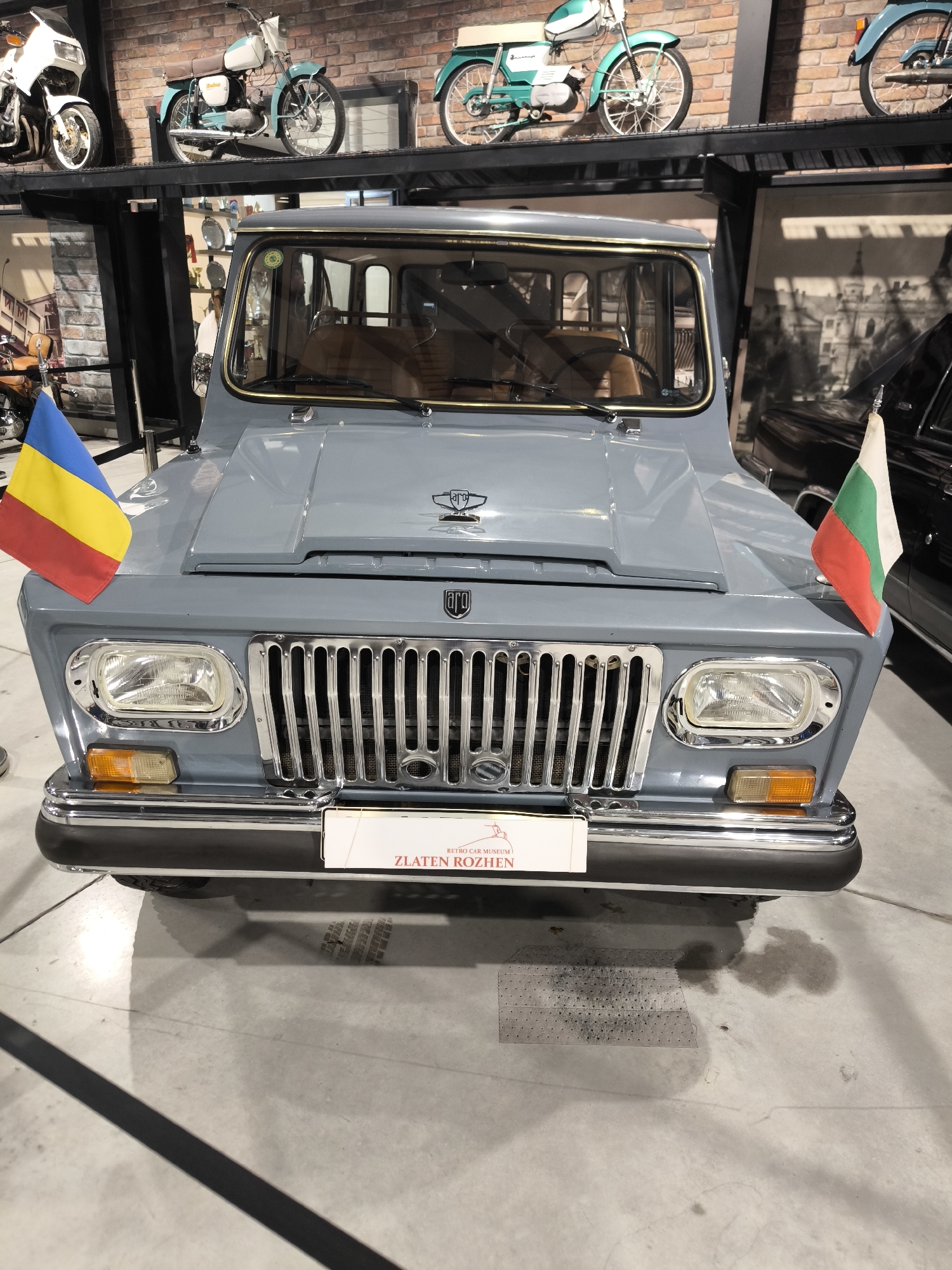 The image shows the front of a light blue ARO vehicle with chrome detailing, likely a vintage SUV, on display in what appears to be a museum or exhibition hall. The car has the Romanian flag mounted on its left and the Bulgarian flag on its right. A sign reading "ZLATEN ROZHEN" is positioned on the front bumper. In the background, other vintage motorcycles are displayed on shelves against a brick wall. The location is presumably in Kapatovo, Bulgaria, as the sign on the vehicle suggests a connection to the Zlaten Rozhen Retro Car Museum. The floor is concrete, and the lighting suggests an indoor environment. There are no people visible in the photograph.