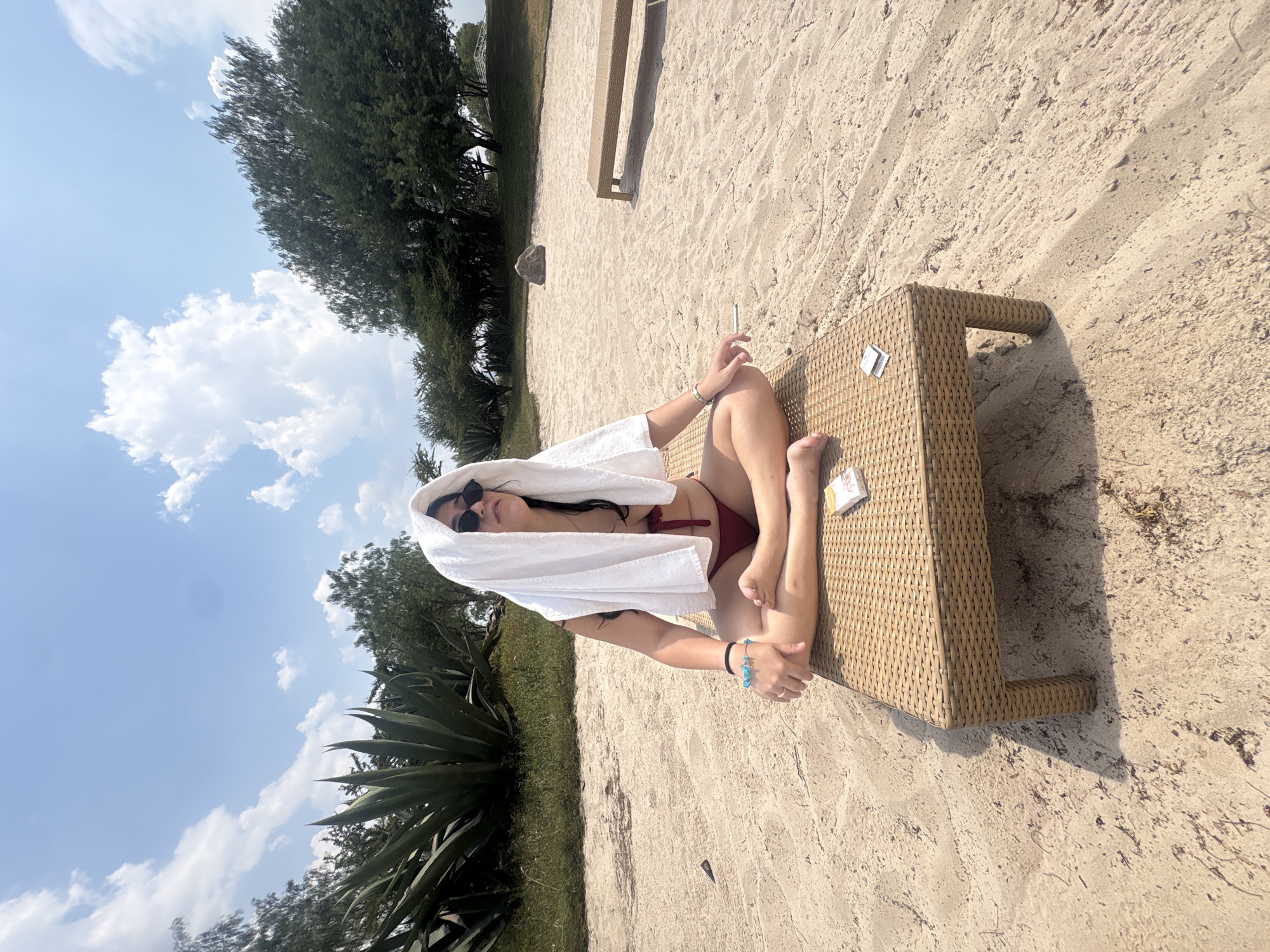 A woman wearing a dark-colored bikini and sunglasses is sitting cross-legged on a wicker-style lounger on a sandy beach. She has a white towel draped over her head. In her right hand, she holds a lit cigarette. On the lounger near her legs are a pack of cigarettes and a lighter. The lounger sits on sand, which transitions into grassy areas with various plants and trees in the background. The sky is blue with scattered white clouds. The location is Pueblo Nuevo de Jasso, Mexico.