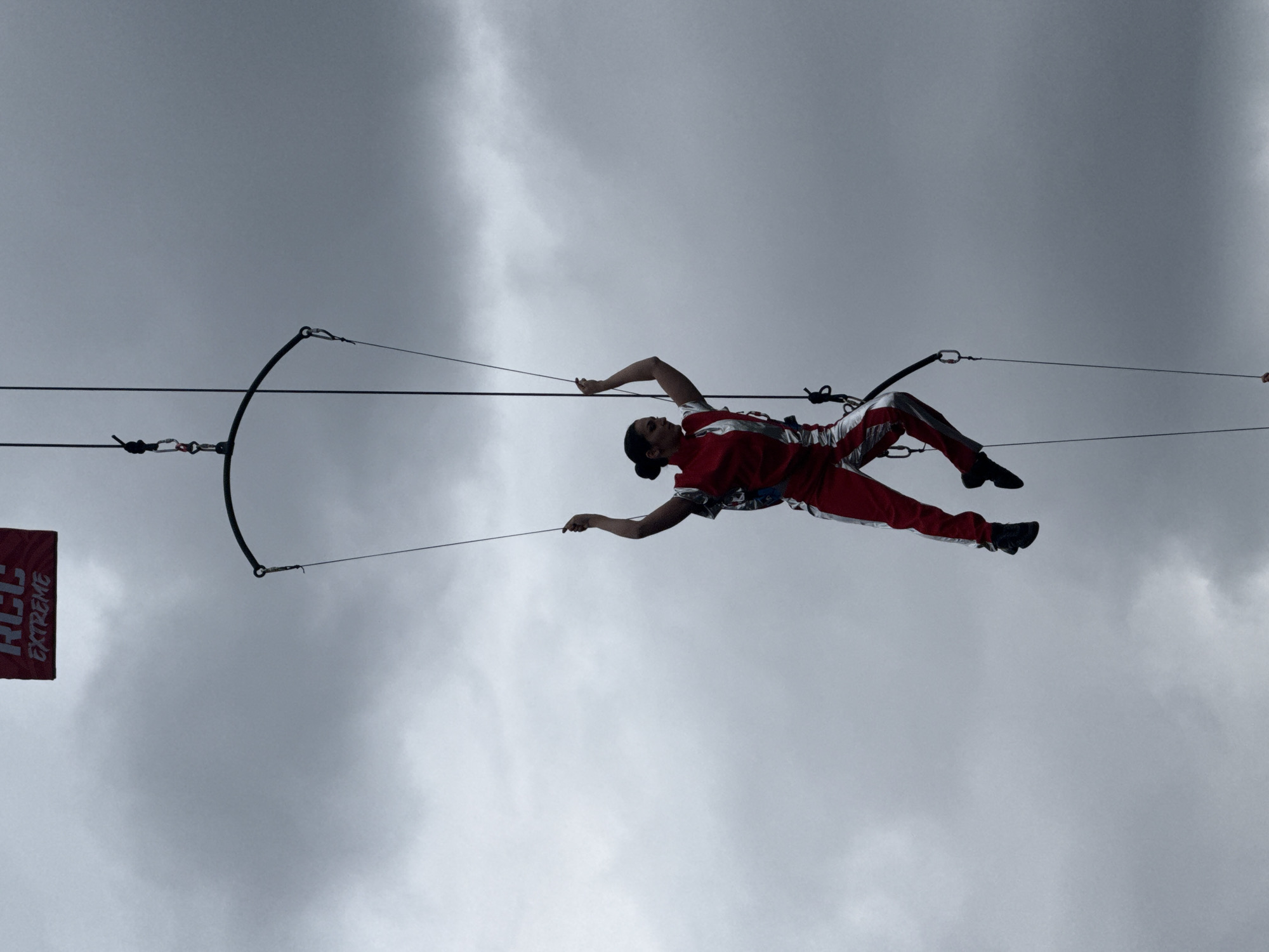 A person in a red and silver jumpsuit is suspended horizontally in the air, attached to a trapeze-like structure, set against a cloudy sky. The person appears to be performing a trapeze act, holding onto the trapeze bar with outstretched arms. A red banner with the words "RCC Extreme" is visible on the left side of the frame. Without additional context, pinpointing the exact location within Yekaterinburg is impossible.