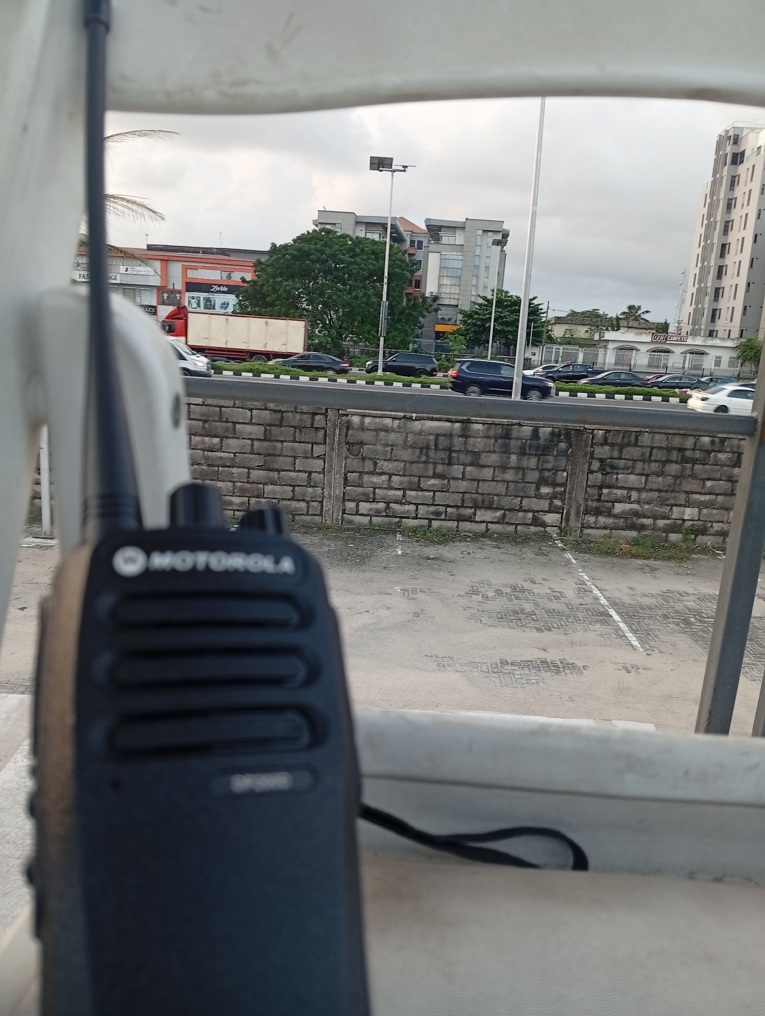 In a city scene, a Motorola walkie-talkie is in the foreground, in sharp focus, with its logo and some buttons visible. Beyond the walkie-talkie, a road with multiple lanes of traffic is visible, separated by a low brick wall. Cars and a large truck are driving on the road. Buildings, including a tall, modern structure and other multi-story buildings, are situated along the road. The sky is overcast, suggesting a cloudy day. The environment appears to be an urban setting with a mix of commercial and residential buildings.
