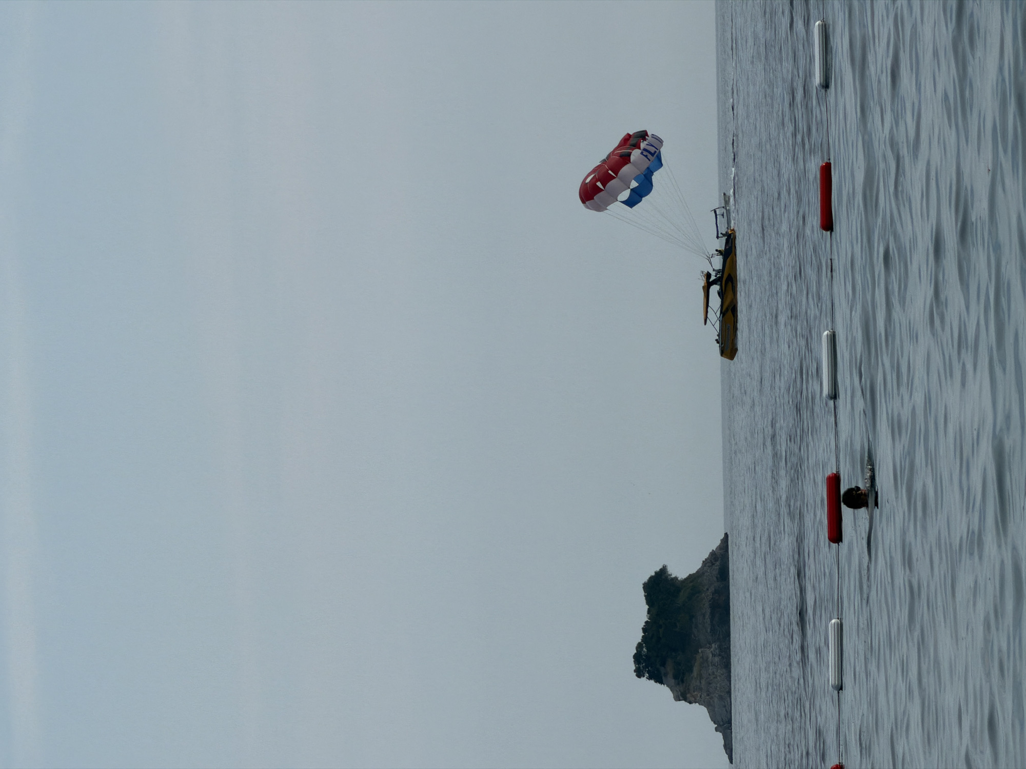 In the city of Budva, Montenegro, a parasailor is seen in mid-air, tethered to a yellow speedboat by strings. The parasail, a colorful red, white, and blue canopy, is inflated against a pale, overcast sky. In the distance, a small, rocky island covered with dense green vegetation rises from the calm, blue water. Closer to the foreground, a person is swimming in the sea, their head and shoulders visible above the surface, wearing sunglasses. Several buoys, colored red and white, mark a boundary in the water. The scene captures a typical recreational activity on a cloudy day in the Adriatic Sea.