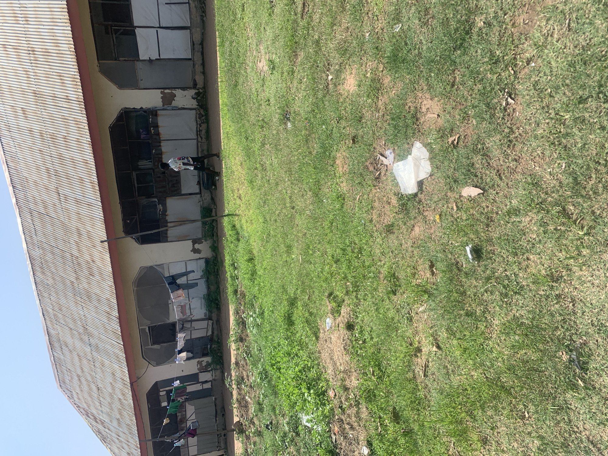 The image depicts the exterior of a building in Jalingo, Nigeria, likely a dormitory or a block of rooms, with a corrugated metal roof and weathered walls. Clothes are hanging on a line in front of some of the windows. A young person, wearing a patterned t-shirt and dark shorts, is walking in front of the building. The foreground is a grassy area with scattered litter, including plastic bags and leaves. The environment appears somewhat neglected, with evidence of debris on the ground. The sky is clear and blue.