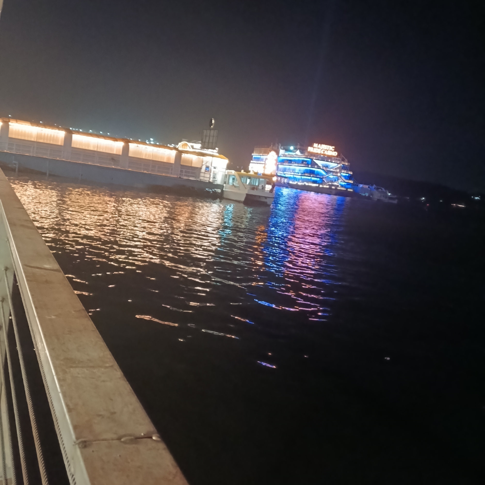 At night in Panaji, India, the image shows a pier extending over water, adjacent to which a small boat is moored. Beyond the pier, a large, multi-decked vessel illuminated with blue and orange lights, identified as the "Majestic Pride Casino," is visible. The water reflects the lights from both the pier and the casino boat. A railing is in the foreground.