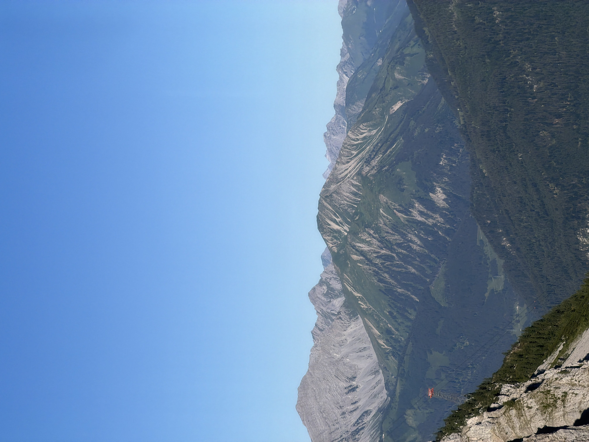 The image depicts a panoramic view of a mountainous landscape under a clear blue sky. The foreground features a steep, rocky cliff face with scattered evergreen trees on its lower slopes. Further into the scene, rolling mountains covered in dense evergreen forests recede into the distance. A red cable car station is visible on the left side of the frame, perched on a mountainside. The overall environment suggests a rugged, natural setting with hiking or tourist activities implied by the presence of the cable car. The location in Grainau, Germany, is a popular tourist destination known for its alpine scenery and outdoor recreation.