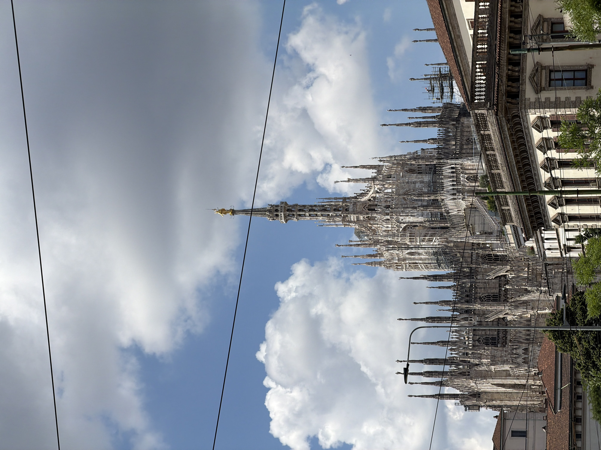 The image presents a striking view of the Milan Cathedral (Duomo di Milano) in Milan, Italy, on a partially cloudy day. The grand Gothic cathedral, constructed from white marble, occupies the majority of the right side of the frame, showcasing its incredibly intricate design. Countless spires, adorned with statues and detailed carvings, rise majestically towards the sky. The highest spire, positioned prominently, is topped with the iconic golden statue of the Madonnina, which stands out against the blue sky.
The sky is a vibrant mix of clear blue and fluffy white cumulus clouds, with some darker, larger cloud formations visible on the left side, adding depth and contrast to the scene. Two thin, dark lines, likely overhead power or tram lines, traverse diagonally across the upper left quadrant of the image.
To the far right, parts of adjacent traditional buildings with windows, balconies, and terracotta roofs are visible, interspersed with green foliage from trees. A street lamp is also present in the lower central part of the frame, near the cathedral structure. Scaffolding can be observed on certain sections of the cathedral, indicating ongoing maintenance or restoration work. The scene is illuminated by natural daylight, suggesting it was captured during the daytime. No people or visible text are present.
