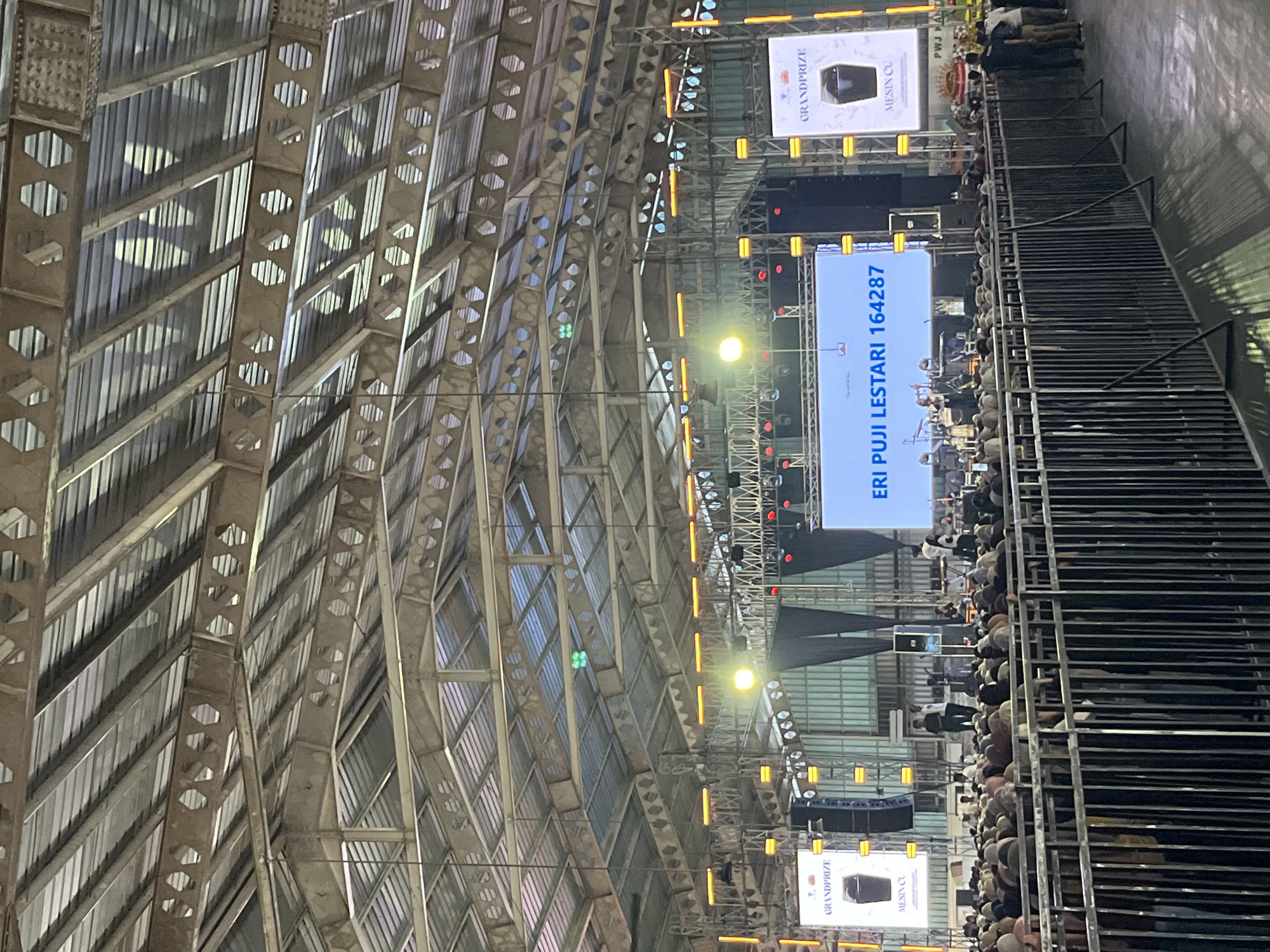 This image depicts an indoor event venue in Pelang, Indonesia, likely a large hall or stadium, characterized by its industrial metal roof structure and extensive lighting rig. A large stage is set up with multiple screens displaying text and graphics. The main screen prominently features the text "ERI PUJI LESTARI 164287" in blue, while side screens show "GRANDPRIZE" and an image of a trash can with the text "MESIN CU". A crowd of people, many wearing white or light-colored head coverings, are gathered in front of the stage, facing it, suggesting they are attending a performance, speech, or religious gathering. They are separated from the stage area by a black metal fence. Several individuals are visible on the stage, some appearing to be performers or organizers, and there is musical equipment present. The overall atmosphere appears to be that of a public event with an audience participating in a communal activity.