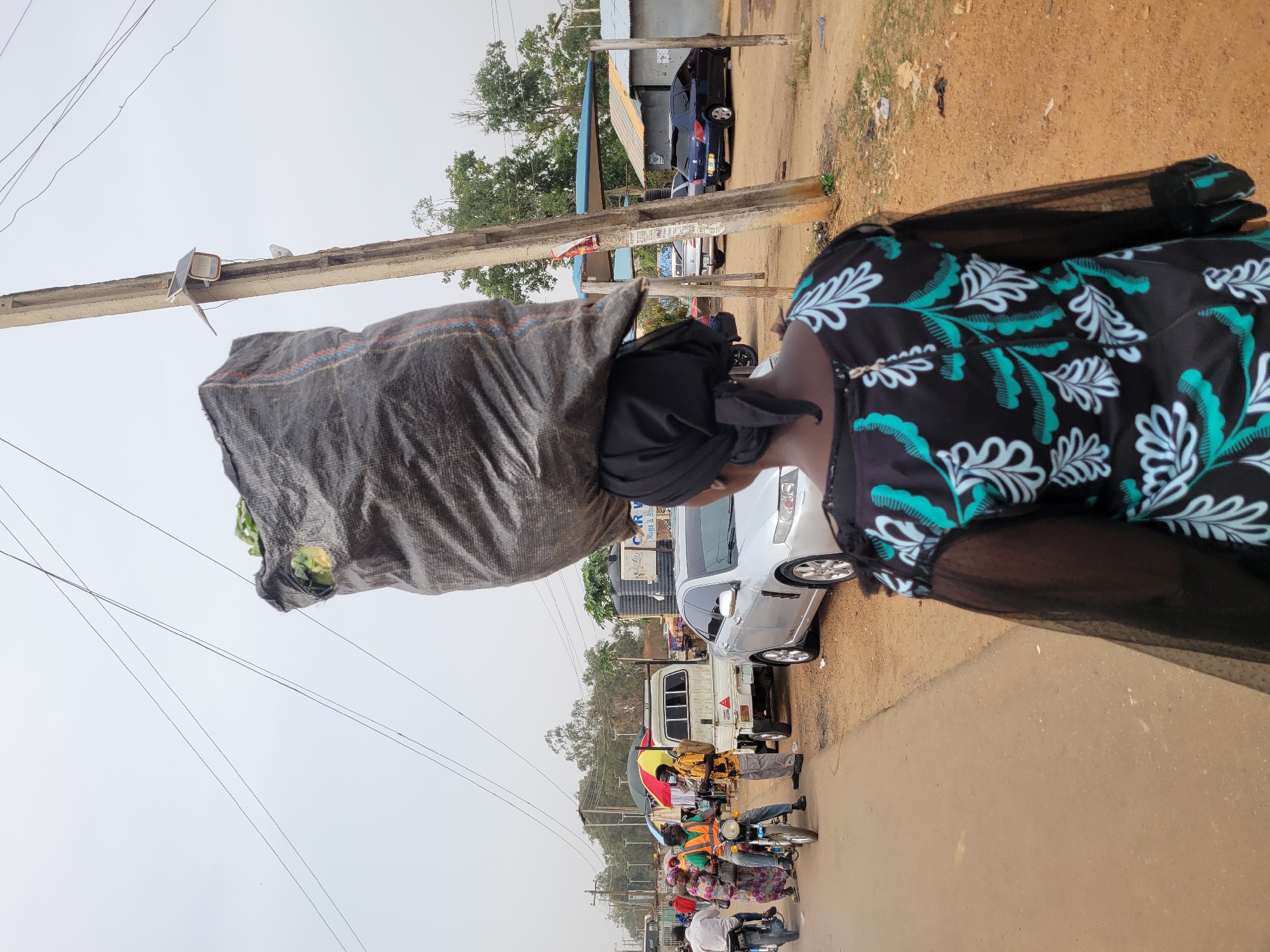 This image depicts a bustling street scene in Jos, Nigeria, centered on a woman expertly balancing a large, heavy sack on her head. The woman, seen from behind and to the side, wears a black headscarf and a black dress with a striking teal and white leafy/floral pattern, featuring sheer black sleeves. The sack, made of a dark, tarp-like material with red and blue stripes, is voluminous and appears to contain green leafy vegetables, some visible at the top.
She walks on the side of a road, which transitions from what appears to be paved asphalt to a dirt shoulder. The sky is overcast, suggesting a daytime scene. In the background, the road is busy with various vehicles, including a white sedan, a blue car, and a white pick-up truck. Numerous people are on motorcycles, many of whom appear to be commercial riders, adding to the vibrancy of the street.
Along the roadside, utility poles with a network of overhead wires are visible. One pole has faded posters or signs attached, but any text is illegible. Buildings and sparse trees line the background, indicative of a semi-urban or developing area. The overall scene captures a typical moment of daily life and commerce, highlighting traditional methods of transport amidst modern traffic.