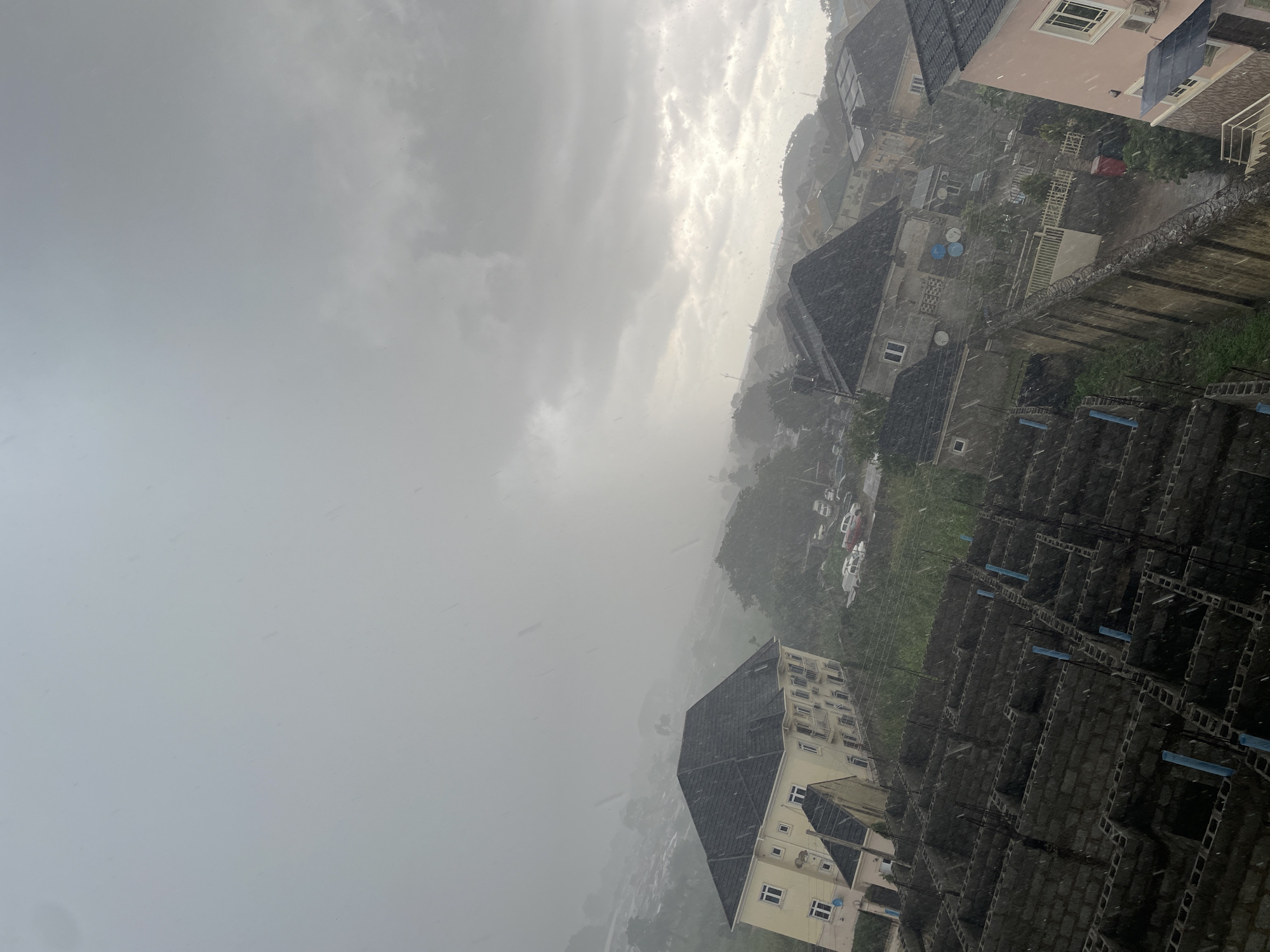 This is an aerial, slightly downward-angled photograph taken during a rainstorm in Abuja, Nigeria. The image depicts a residential area with multiple buildings and a construction site. In the foreground, a partially constructed building made of grey cinder blocks is visible, with blue pipes protruding from its walls. To the left, a large, cream-colored, multi-story building with a dark roof is prominent. Further into the scene, other houses with varying roof types and architectural styles are scattered amongst lush green vegetation and trees. The sky is heavily overcast with grey clouds, and visible raindrops streak across the frame, indicating active rainfall. A few cars are parked in the distance. The overall impression is of a cloudy, rainy day in an urban or suburban setting.