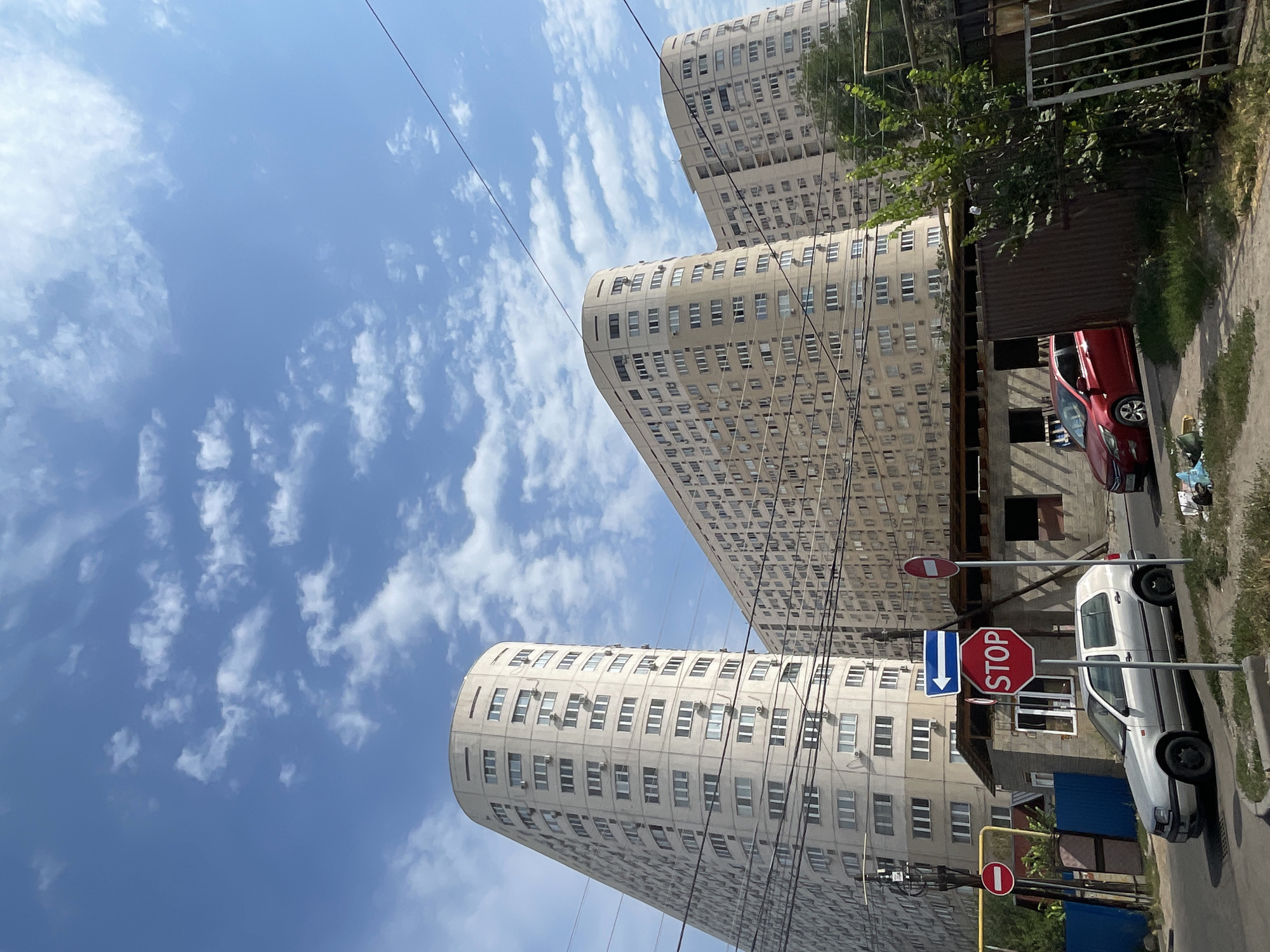 In Almaty, Kazakhstan, a street scene unfolds under a bright blue sky with scattered white clouds. The foreground features a gray sedan parked on an asphalt road, its wheels slightly turned. Beside it, a red hatchback is partially visible. A small, utilitarian building with a dark roof stands behind them, with construction materials and debris scattered nearby. Multiple traffic signs are present, including a red octagonal "STOP" sign, a blue sign with a white arrow pointing left, and a circular red and white prohibition sign.
Dominating the background are three tall, beige apartment buildings with a distinct curved architectural style. These structures are densely populated with windows and air conditioning units, indicative of a residential area. Overhead, power lines crisscross the sky, adding to the urban feel of the scene. Green trees and a metal fence are also visible on the right side of the image. The overall impression is one of a typical urban environment in Kazakhstan.