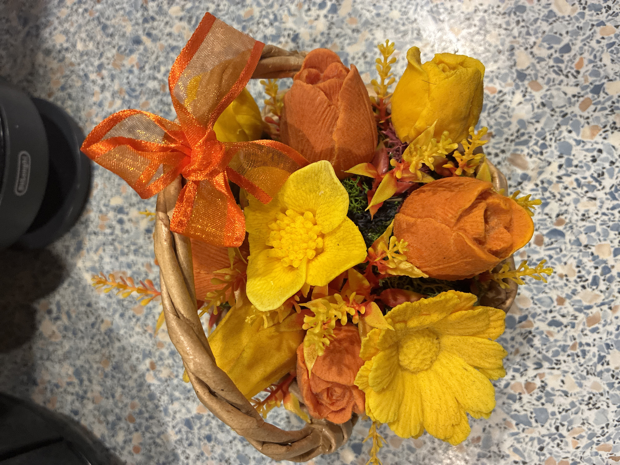 An orange sheer ribbon bow is tied to the handle of a small woven basket filled with artificial flowers. The flowers are in shades of yellow and orange, with some small yellow and red accents. The basket is placed on a speckled countertop.