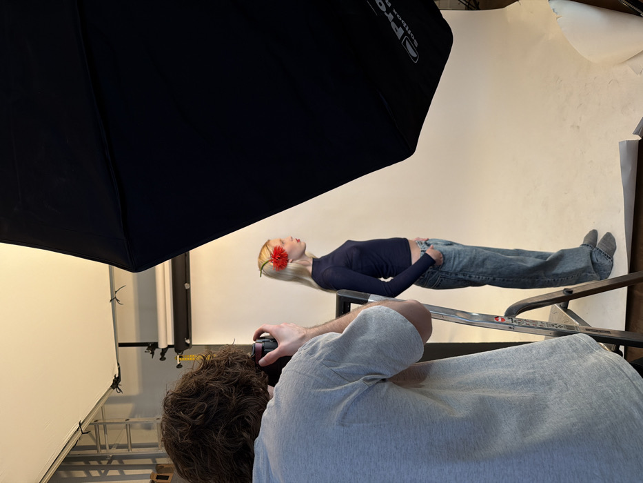 An indoor photography studio session is depicted, purportedly located in Paris, France. A male photographer, viewed from behind, with dark curly hair and wearing a grey short-sleeved t-shirt, is positioned on the bottom step of a silver-colored folding ladder. He holds a dark camera to his eye, actively engaged in photography. To his right, a female model stands in profile, facing right, against a seamless white paper backdrop. She has long blonde hair with a red flower accessory behind her left ear and is wearing red lipstick. Her attire consists of a fitted navy blue long-sleeved top, light blue denim jeans with hands in pockets, and grey socks. A large, black Profoto-branded softbox on a stand partially fills the right side of the frame, oriented towards the model. Studio lighting equipment and support structures are visible in the background and overhead. The floor is dark brown.