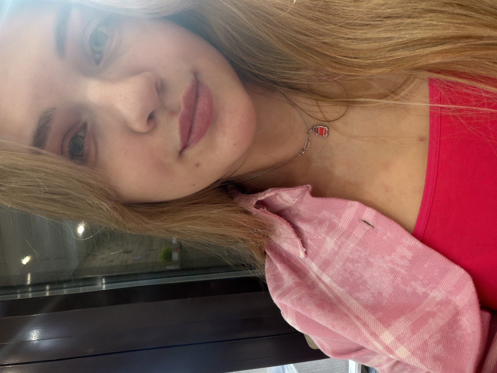 A young woman is pictured in a close-up, slightly angled shot. Her hair is light brown, and she is wearing a pink plaid shirt over a bright pink top. A necklace with a small, red pendant is visible around her neck. The background is out of focus, but suggests an indoor setting, possibly near a window with dark framing. The location within Naberezhnye Chelny, Russia is impossible to determine from the provided image.
