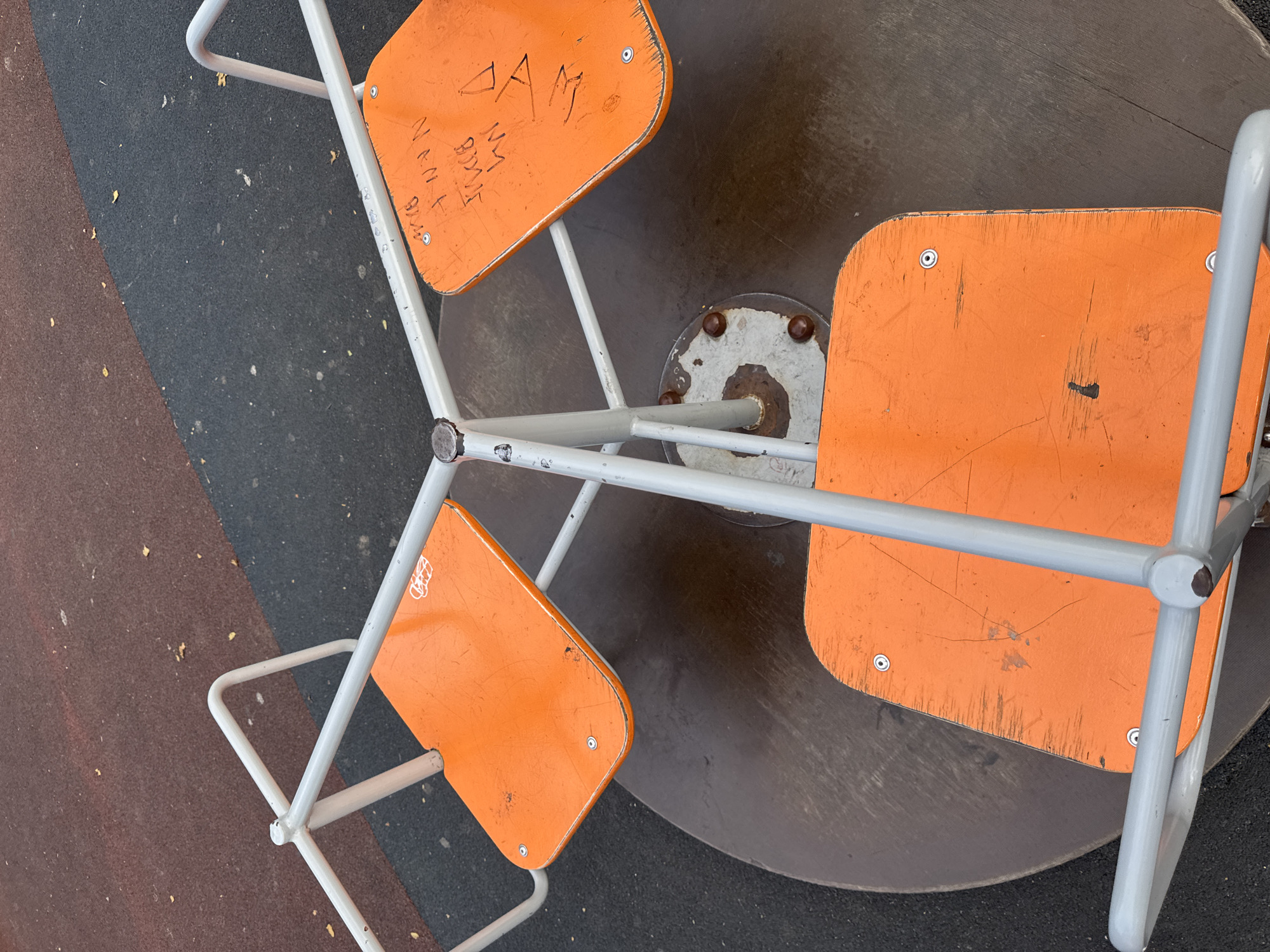 A top-down, close-up shot shows an orange merry-go-round on a playground in Verkhnerussky, Russia. The merry-go-round has three orange seats, each with a grey metal bar for support. The seats appear worn with scratches and some graffiti. The ground around the merry-go-round is a mix of dark grey and reddish-brown rubberized playground surfacing, with a few fallen leaves visible. The merry-go-round itself is made of grey metal.