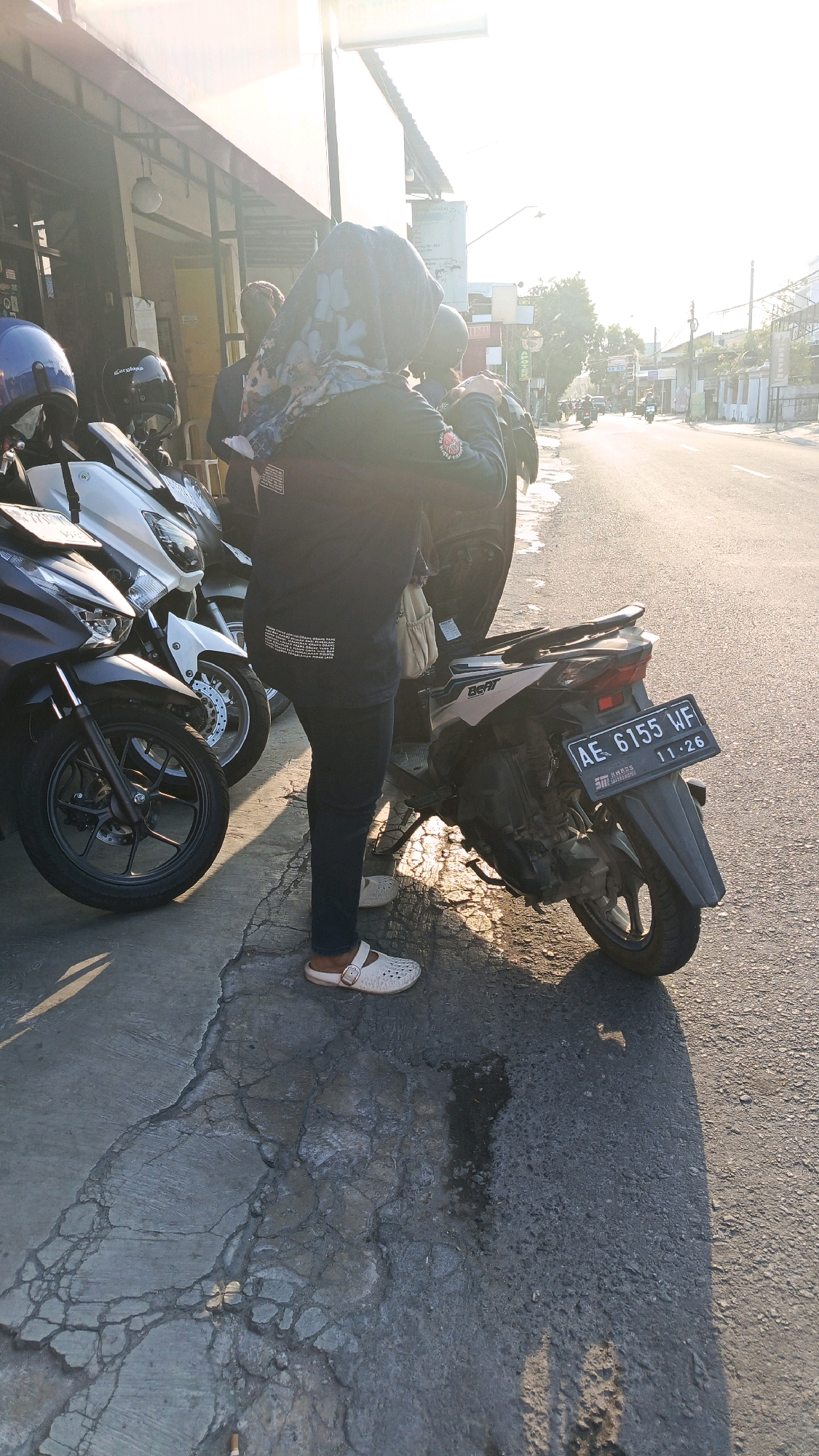 A person wearing a dark hijab and dark pants stands next to a motorcycle parked on the side of a street in Ronowijayan, Indonesia. The person is wearing white slip-on sandals. The motorcycle has a license plate that reads "AE 6155 WF 11-26". Other motorcycles are parked nearby. The street appears to be in a commercial area, with buildings and other vehicles visible in the background. The sun is shining, casting shadows on the ground. The pavement is cracked and uneven.