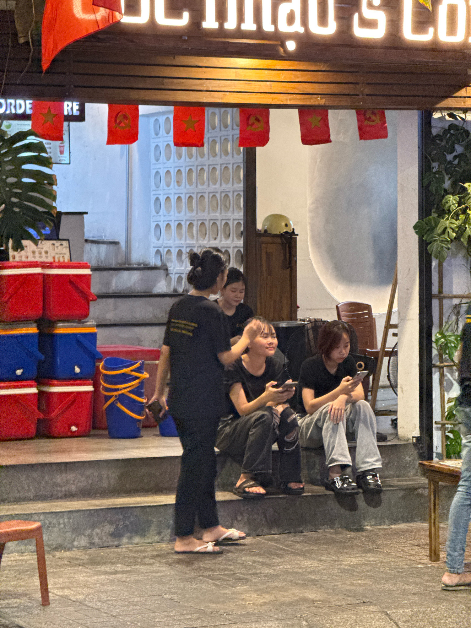 The image depicts a street-level view of a cafe in Nha Trang, Vietnam, likely in the evening. The cafe's facade features a prominent neon sign with text that is partially visible, reading "u hau's Co...". Below the sign, a wooden overhang displays several small red flags, some bearing the hammer and sickle symbol, and others a yellow star on a red background, suggestive of Vietnamese national or political symbolism.
In the foreground, a woman wearing a black t-shirt and dark pants stands on a set of concrete steps, holding a dark-colored mobile phone. She is facing towards the right side of the frame, with her body angled away from the viewer. To her right, three young women are seated on the same steps. The woman closest to the viewer is wearing a black t-shirt and dark, possibly ripped, jeans. She is holding a mobile phone in her left hand and appears to be looking at it. Next to her, a woman in a black t-shirt and light grey pants is also looking down at a mobile phone in her lap. To her right, another woman in a light grey t-shirt and dark pants is seated, looking towards the left and touching her head with her right hand.
To the left of the steps, a stack of red and blue plastic coolers is visible. A blue bucket with orange handles is placed on the steps. In the background, a pale-colored wall with a patterned texture is visible, as well as a wooden cabinet with a yellow helmet on top. Green foliage is present on the right side of the frame, indicating an outdoor seating area or a plant display. The ground in front of the steps is paved with square tiles. The lighting suggests an evening or nighttime setting, with artificial light illuminating the scene.
