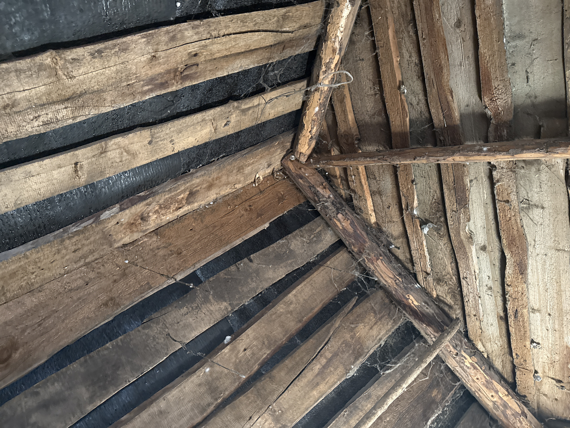 This is a close-up, low-angle photograph of the wooden interior of a rustic structure, possibly a barn or shed, located in Чуй-Атасево, Russia. The scene is dominated by rough-hewn wooden planks, oriented diagonally and horizontally, forming a roof structure. Some planks are weathered and cracked, revealing their age and exposure to the elements. Dark, possibly tarred, material is visible between some of the diagonal planks, suggesting a weatherproofing layer. Cobwebs are present, clinging to the wooden beams and rafters, indicating a lack of recent human activity. No people or animals are visible. The lighting is dim, and the overall impression is one of disuse and age.