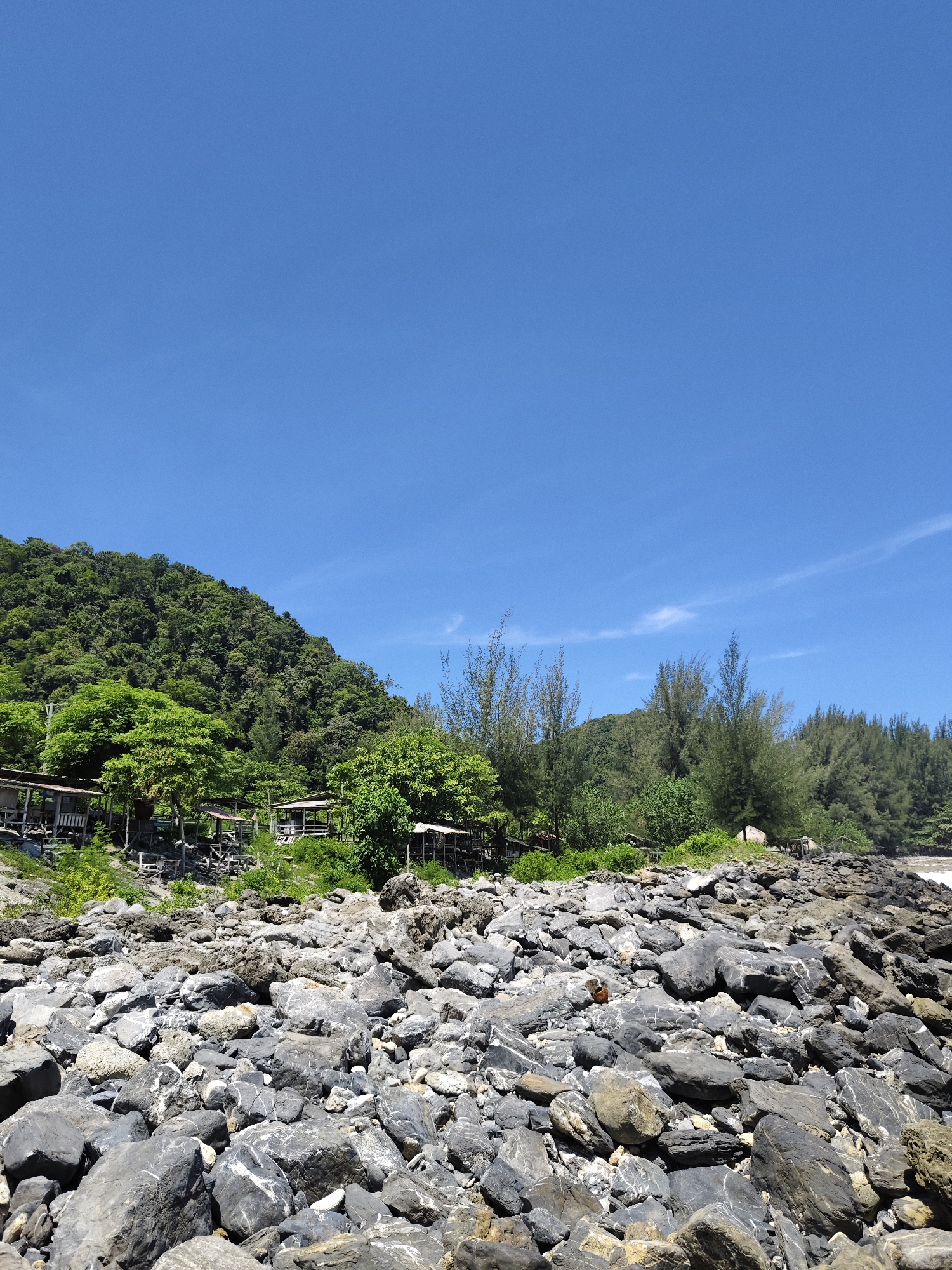 The image depicts a rocky shoreline with a lush, green hillside in the background. The foreground is dominated by a dense collection of dark grey and black rocks of various sizes, scattered across the ground. Some of these rocks exhibit white veining. Beyond the rocks, there is a band of vegetation including trees and bushes. Further back, several small, simple wooden structures with weathered roofs are nestled among the trees, suggesting a small settlement or resort. The hillside is covered in dense foliage, creating a green, textured landscape that rises towards the clear blue sky. The sky itself is a vibrant blue with a few wispy white clouds. The overall scene suggests a natural coastal environment in Naga Umbang, Indonesia, characterized by its rocky terrain, abundant greenery, and the presence of rudimentary structures. There are no people visible in the image.