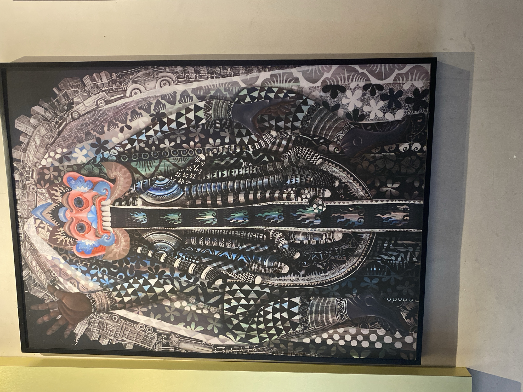 A framed art piece hangs on a wall, the bottom of which is positioned on a green ledge. The artwork is a complex, graphic piece dominated by black, white, and gray tones. A vividly colored mask-like face in shades of red, orange, and blue is positioned on the left side of the piece. The artwork is composed of various patterns, including floral designs, geometric shapes, and images of cars and buildings. An image of a hand is also visible. The scene is in a room in the city of Mengwi, Indonesia.