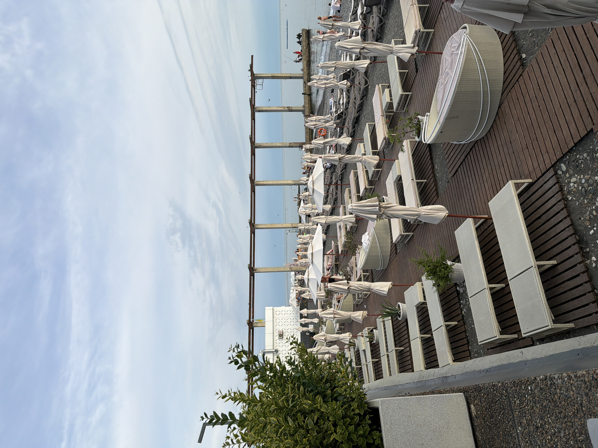 The image depicts an outdoor beach scene, likely in Sochi, Russia, given the style of the resort and the presence of the Black Sea. A wide expanse of lounge chairs and closed umbrellas covers a pebble beach, with wooden walkways interspersed between them. In the foreground, a large planter with a leafy green bush is visible on the left, and several rows of white lounge chairs with light-colored cushions are neatly arranged. Further back, more lounge chairs and umbrellas are visible, some with people seated or reclining on them. A distinctive, multi-arched concrete structure extends over the water in the background, possibly a pier or part of a former structure. Several individuals are present on the pier, some appearing to be fishing. The sky is partly cloudy with a hazy appearance, suggesting overcast or dusk conditions. The overall atmosphere is one of a quiet, well-maintained resort beach.