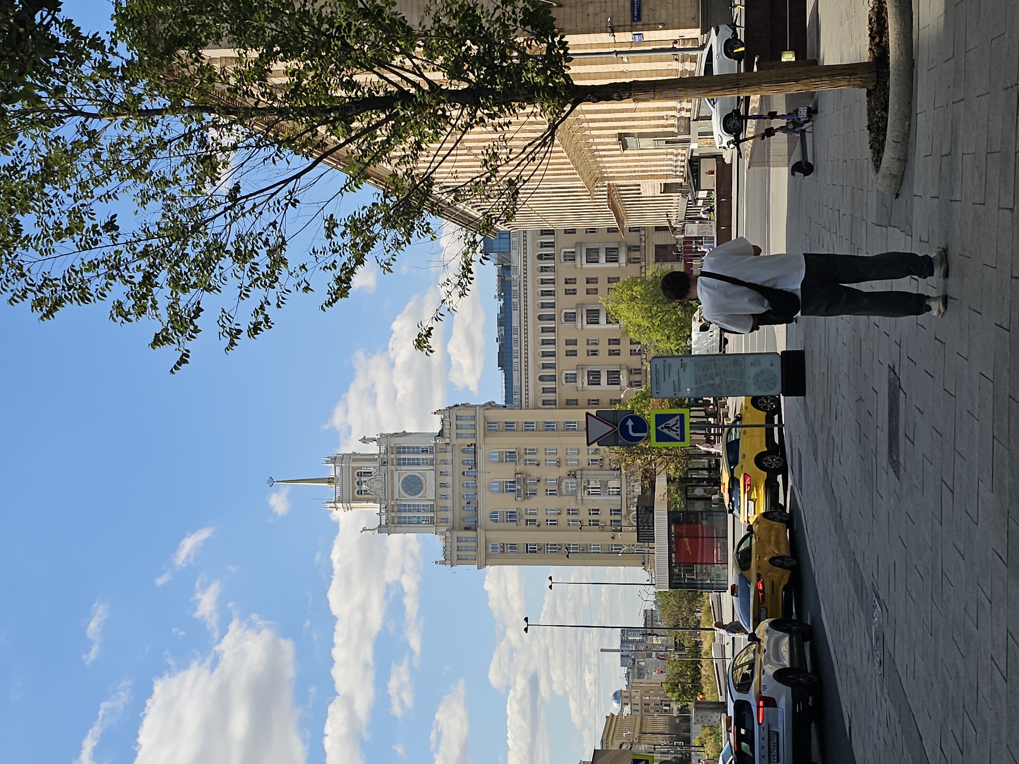 In a Moscow street scene, a person stands on a sidewalk, facing away from the camera. They are wearing a white t-shirt and dark pants, with a dark bag slung over their shoulder. In the background, a tall, pale yellow building with a prominent clock tower dominates the skyline. The building features a golden spire and decorative elements. A line of yellow cars, likely taxis, is parked or moving on the street in front of the building, with a white SUV further down the road. The sidewalk is paved with grey tiles. A young tree grows on the right side of the frame, obscuring part of an adjacent building with a classical facade. The sky is blue with scattered white clouds. Traffic signs and streetlights are visible along the street. The overall impression is a sunny day in an urban environment.