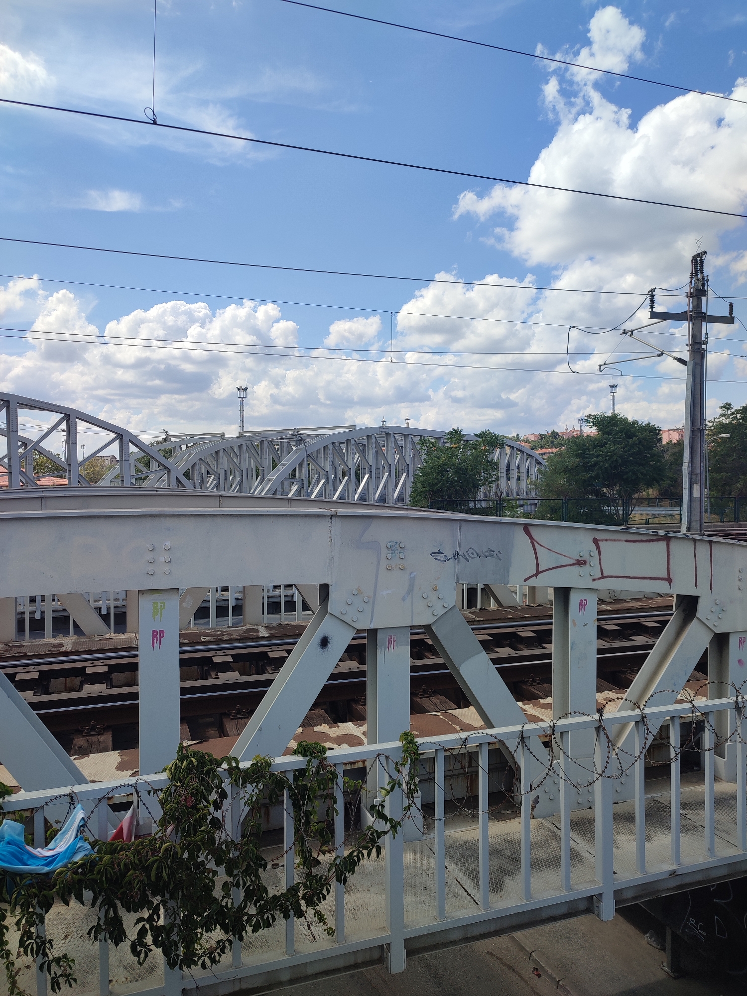 The image shows a view of railway tracks and bridges under a bright blue sky with white clouds. In the foreground, there is a light-colored bridge with visible bolts and some graffiti. Barbed wire and some greenery are attached to the railing of this bridge. Beyond this bridge, another, more distant, light-colored truss bridge is visible. The railway tracks are laid out between the supports of the foreground bridge. In the background, trees and buildings can be seen, along with a utility pole with electrical wires overhead. The overall scene depicts an urban infrastructure environment with a railway line and bridges. The image appears to be taken from a slightly elevated position, looking across the railway tracks.